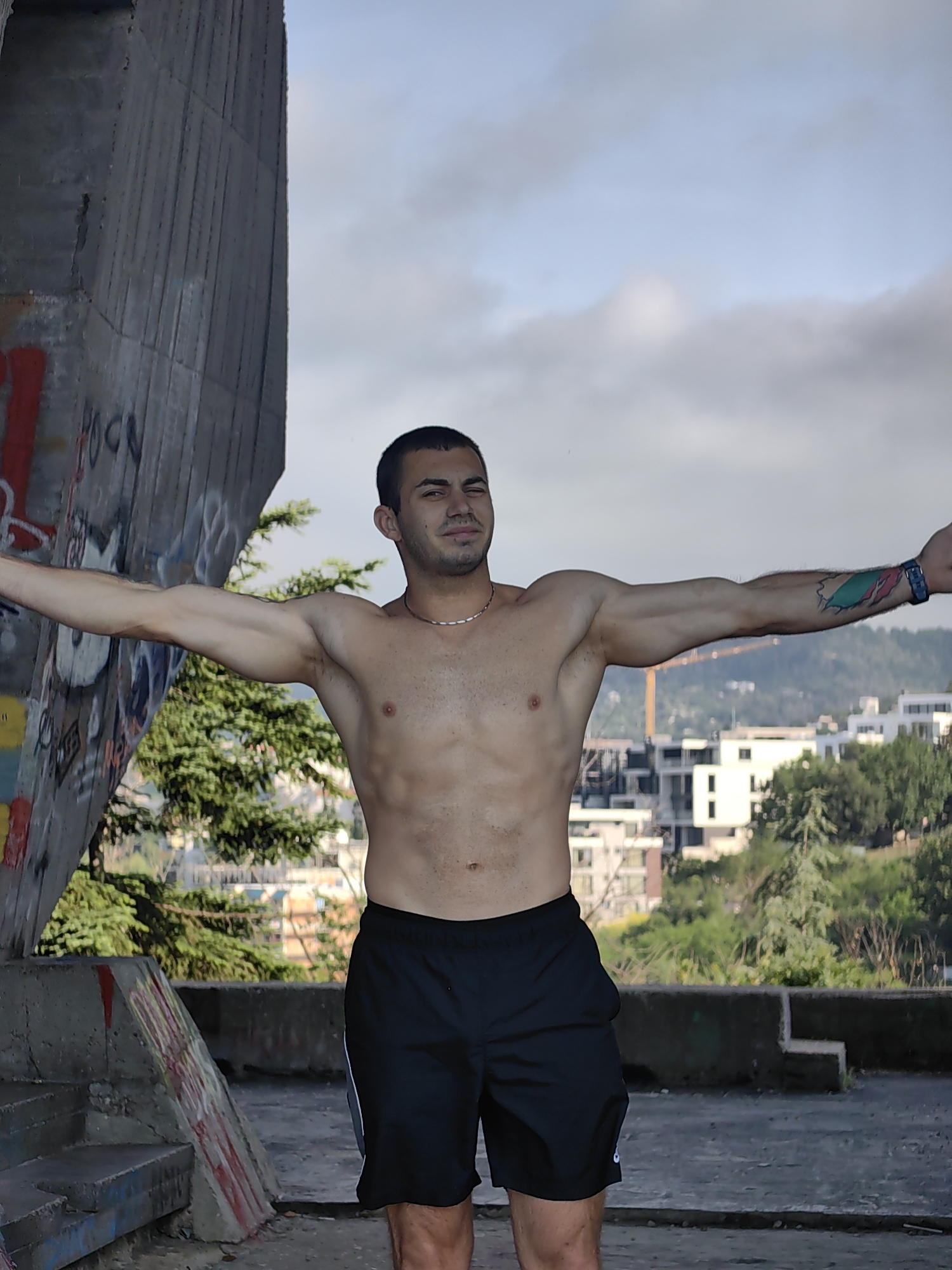 A young man with a muscular physique stands with his arms outstretched, leaning against a large, graffiti-covered concrete structure on a rooftop overlooking the city of Varna, Bulgaria. The man is wearing black athletic shorts. He has short dark hair and a thin necklace. A colorful tattoo is visible on his right arm. In the background, the city's urban landscape unfolds, with buildings and greenery under a cloudy sky. The concrete structure is weathered and shows signs of graffiti art, including red and black markings. Stairs and a concrete railing are visible in the foreground. The scene appears to be set in an outdoor, elevated location, possibly an abandoned or partially constructed building, offering a panoramic view of the city.