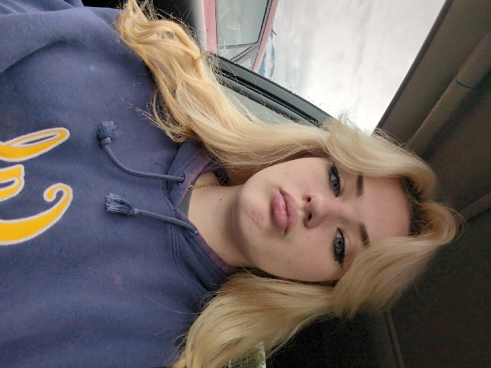 A young blonde female is depicted from the chest up, seated inside a vehicle. She has fair skin, blue eyes accentuated with black winged eyeliner, and light pink lips pursed into a pout. Her long, wavy blonde hair is styled with face-framing layers and volume at the crown. She wears a navy blue hooded sweatshirt with a partially visible yellow and white graphic at the bottom, a grey undershirt, and a thin gold chain necklace. Faint reddish-purple discoloration is present on the hoodie around the neckline. Through the vehicle's window on the left, an overcast grey sky and distant snow-capped mountains are visible, consistent with the mountainous geography of Area D (Elaho/Garibaldi), Canada. The roof of a red vehicle is partially visible outside, and raindrops or condensation are present on the window glass. The subject looks directly towards the camera.