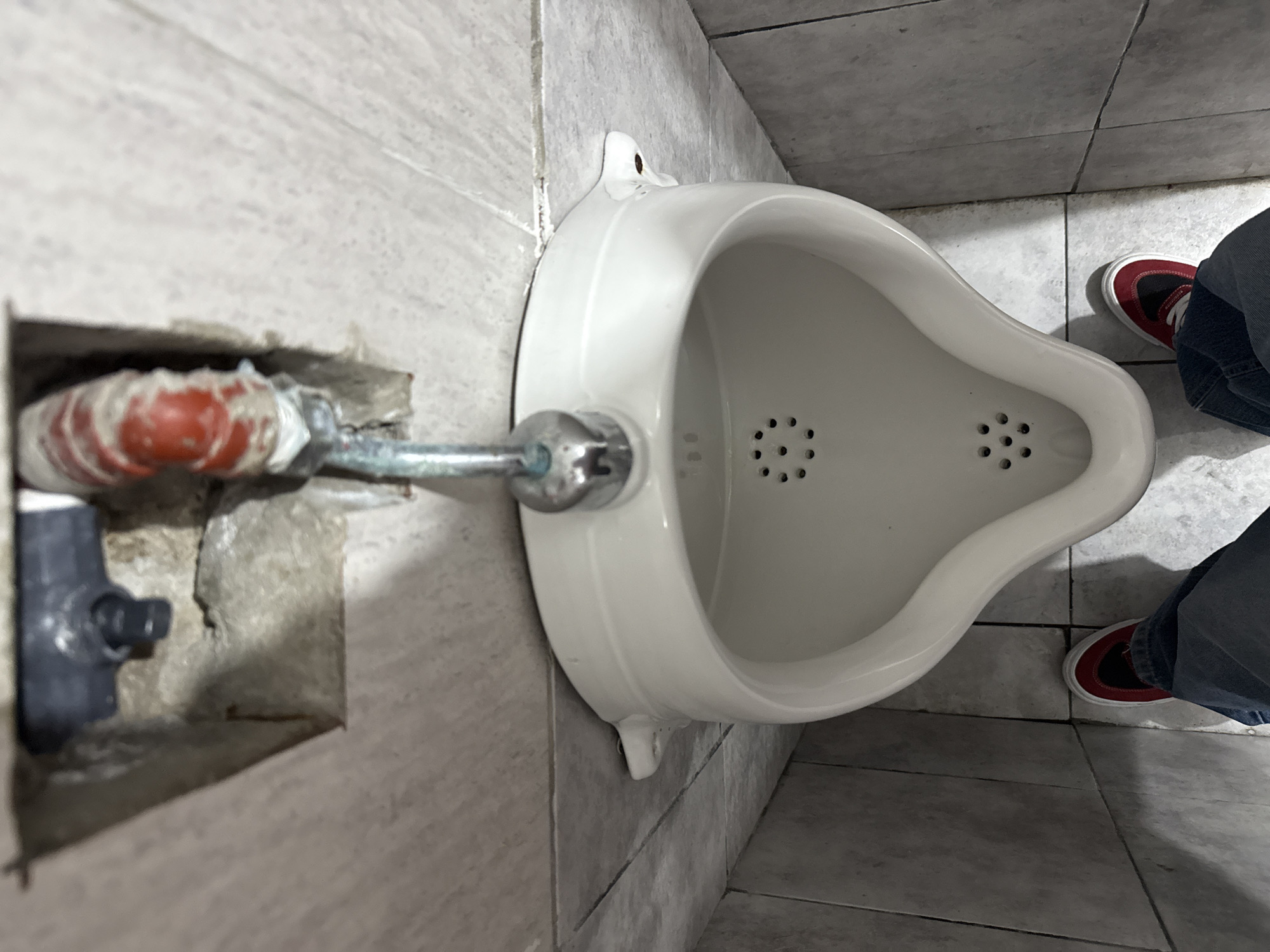 An overhead shot displays a white urinal fixed to a tiled wall, with visible plumbing. A person wearing jeans and red sneakers stands to the side. The scene is likely inside a public restroom with tiled floors and walls. The location cannot be determined with this image.
