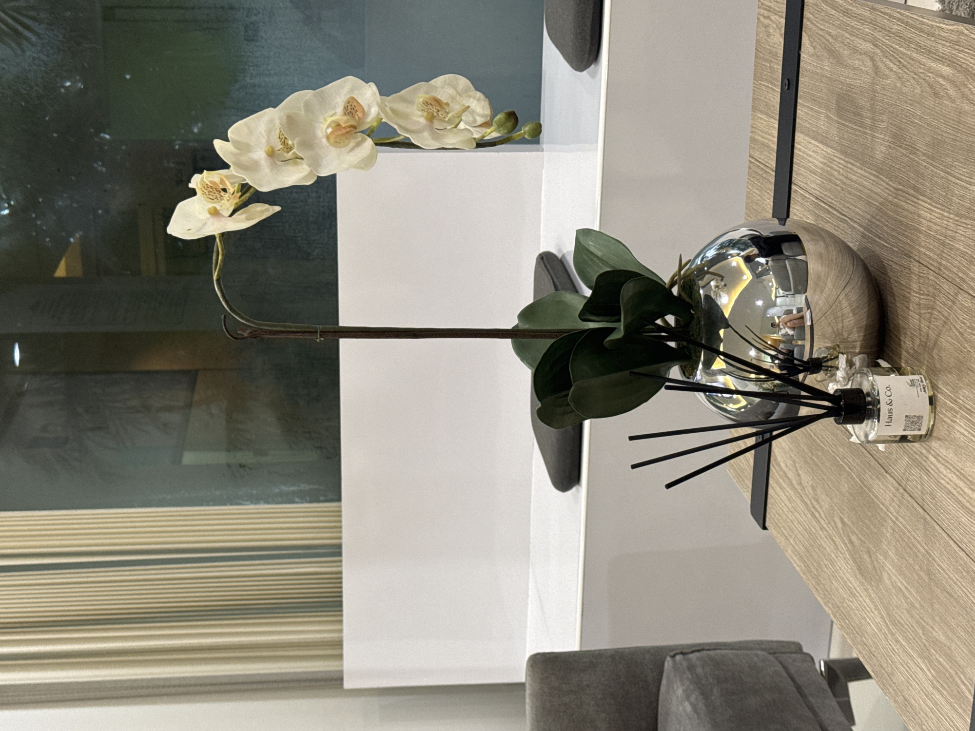 This image captures a still life arrangement on a light wood table, likely in a modern indoor setting. The primary subjects are a decorative artificial orchid with creamy white petals and yellow-speckled centers, and a shiny, spherical metallic vase. Dark green leaves, appearing to be part of the orchid arrangement, spill out from the vase.
Beside the vase sits a small glass bottle of diffuser reeds, with the label clearly visible. The text on the label reads "Haus & Co." followed by some smaller text that appears to be a scent name, possibly "Olive". The diffuser reeds are black and arranged in the bottle.
The background is a mix of elements. To the left, a large window or glass surface reflects a muted, possibly outdoor scene with trees and indoor lighting. Below the window, horizontal blinds or a slatted surface creates a striped pattern. To the right of the arrangement, a white, minimalist surface, possibly a wall or a shelf, forms a clean backdrop, with a dark, thin vertical element possibly indicating a fixture or edge. A sliver of a grey upholstered piece of furniture is visible in the lower foreground, suggesting a living space.
The lighting appears to be indoor ambient light, possibly supplemented by natural light from the window, creating soft reflections on the metallic vase. There is no obvious activity or event taking place; the scene is static and decorative. No specific time of day or weather conditions can be inferred beyond the indoor setting. The location context of Chiang Mai City Municipality, Thailand, is not directly evident from the visual cues in the image itself.