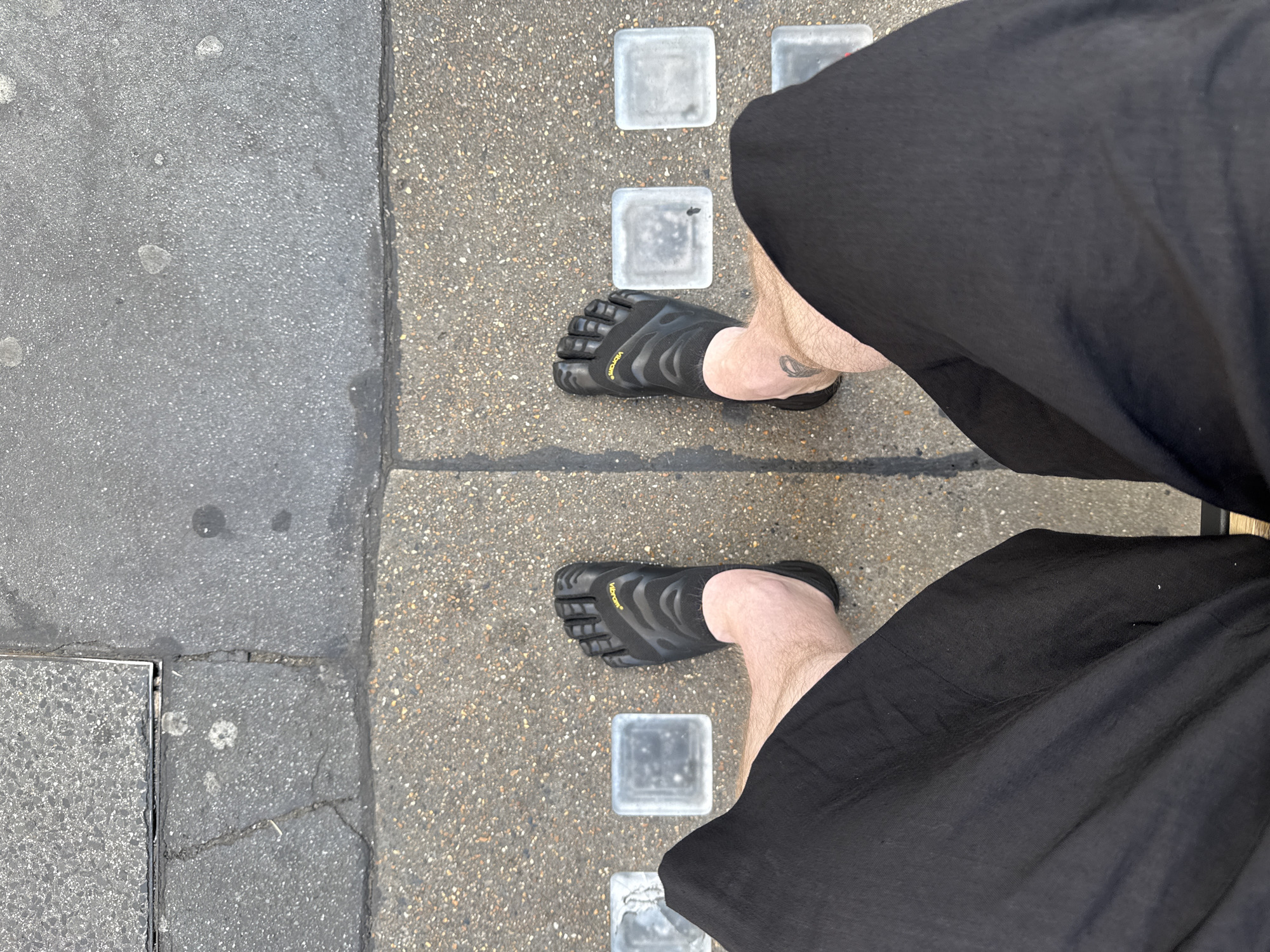 A first-person view shows a person standing on a sidewalk in London, United Kingdom. The person is wearing black toe-separation shoes and loose black pants. The sidewalk is made of gray concrete with a strip of smaller gravel-like concrete. A row of square glass blocks is embedded in the gravel strip.