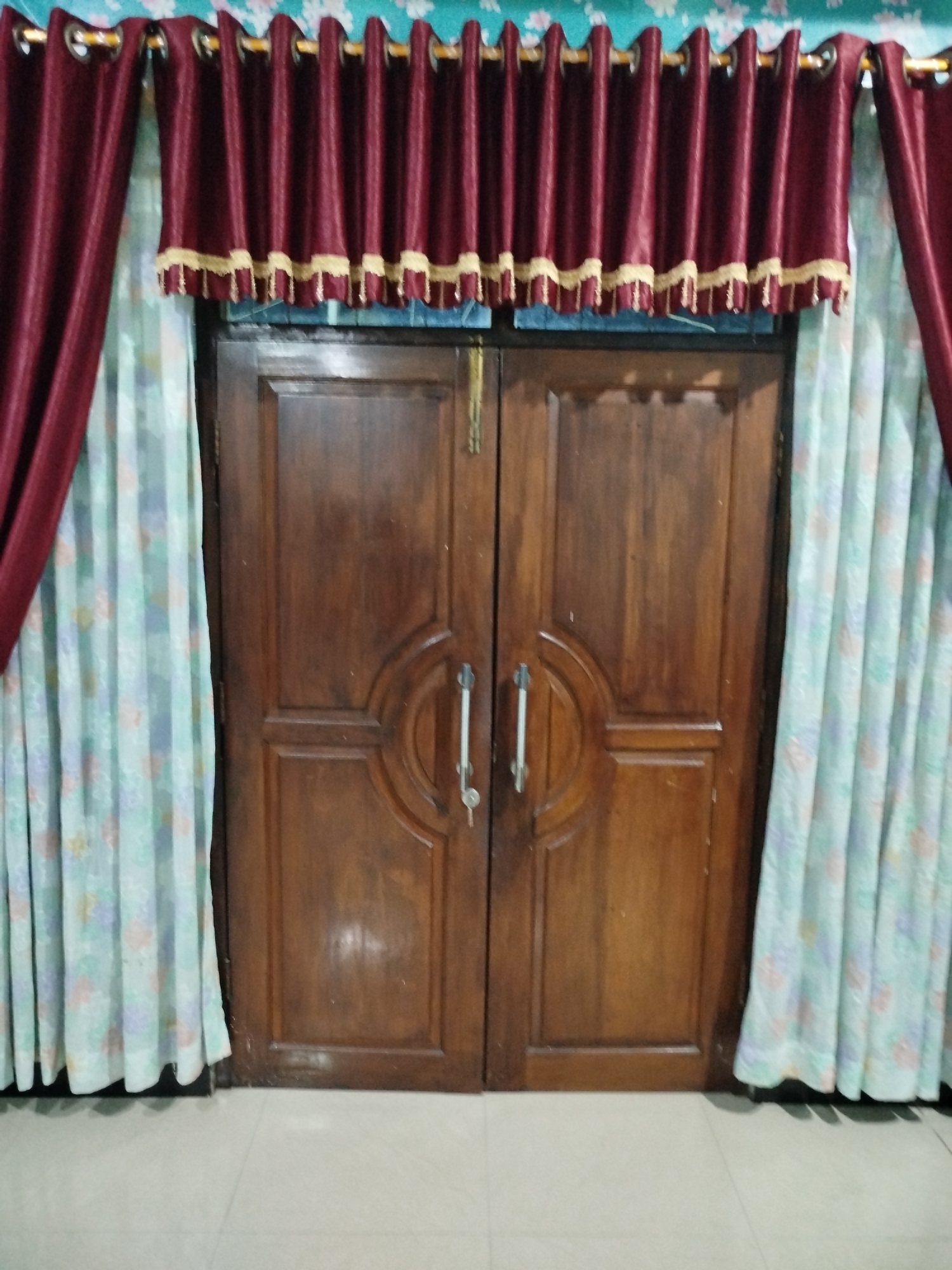 The image displays a wooden double door with ornate carvings and two silver handles. A key is visible in the lock of the right door. The door is flanked by light blue curtains with a floral pattern, and a burgundy curtain with gold trim hangs above the door. The flooring appears to be tiled. The setting is indoors, likely a residential interior, and the scene is static with no visible people or actions. The image is a straightforward depiction of a doorway, devoid of any specific context that would place it in a particular city or region.