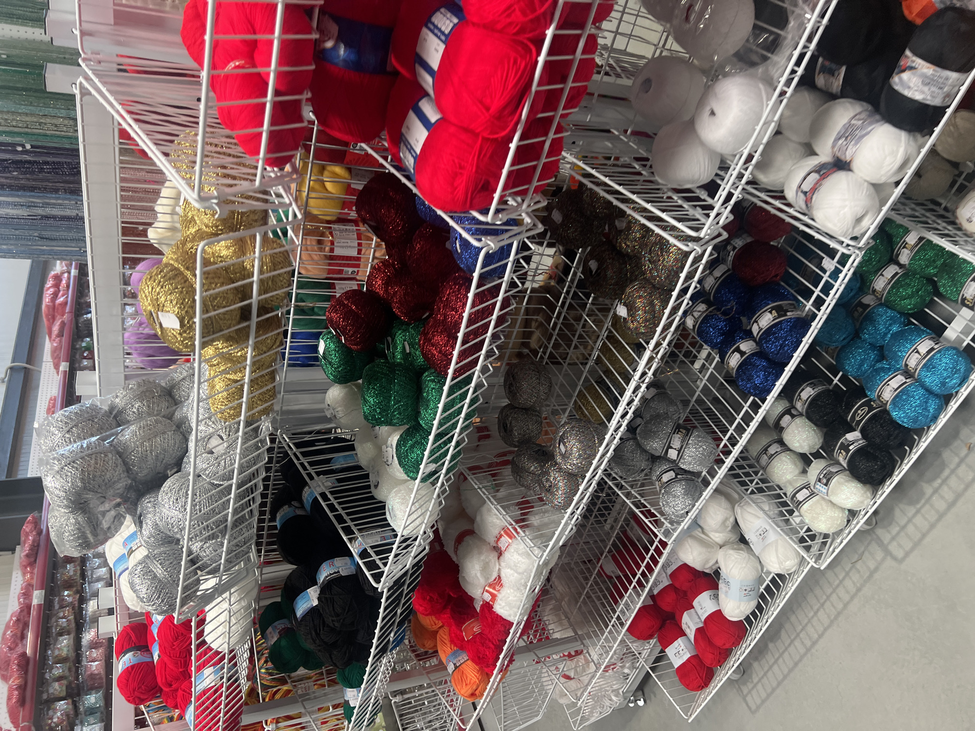 The image shows a close-up view of shelves filled with yarn in various colors. The yarn is neatly arranged in white wire baskets. Colors visible include red, gold, silver, black, white, green, blue and brown. The shelves appear to be part of a retail display in a shop, possibly located in Burgas, Bulgaria. There are no people visible in the image.