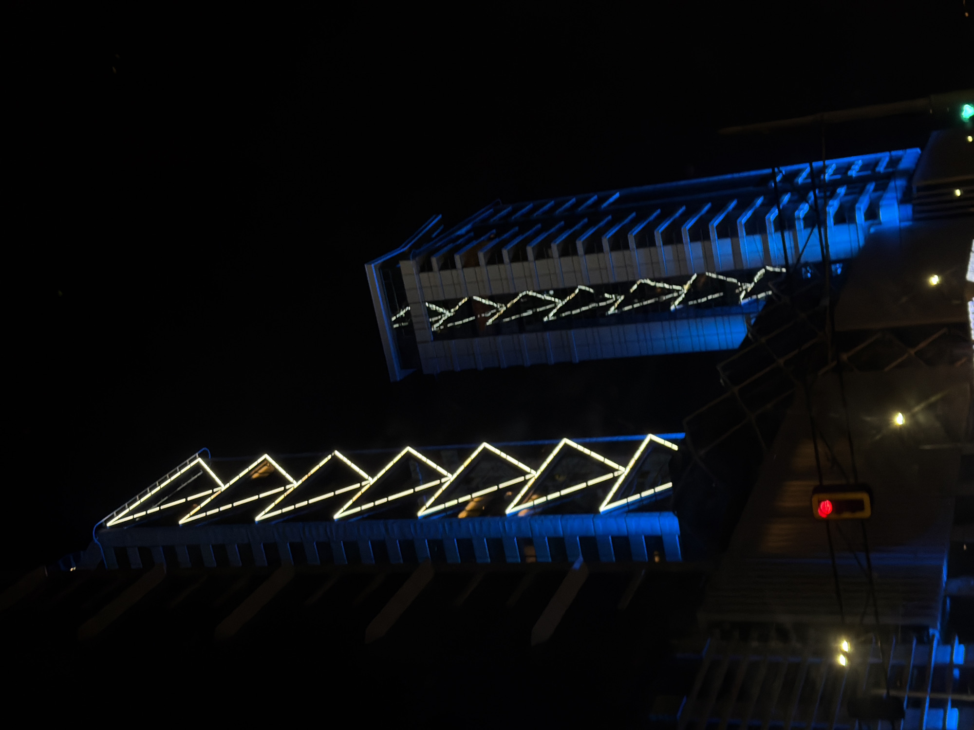 The image depicts a nighttime view of buildings in Sofia, Bulgaria. The buildings are illuminated with blue light, and yellow lights are arranged in a triangular pattern along the top of one structure. The lower structure has long supports extending down. In the foreground, what appears to be scaffolding with exposed bulbs can be seen, along with a red traffic light. The background is a dark night sky.