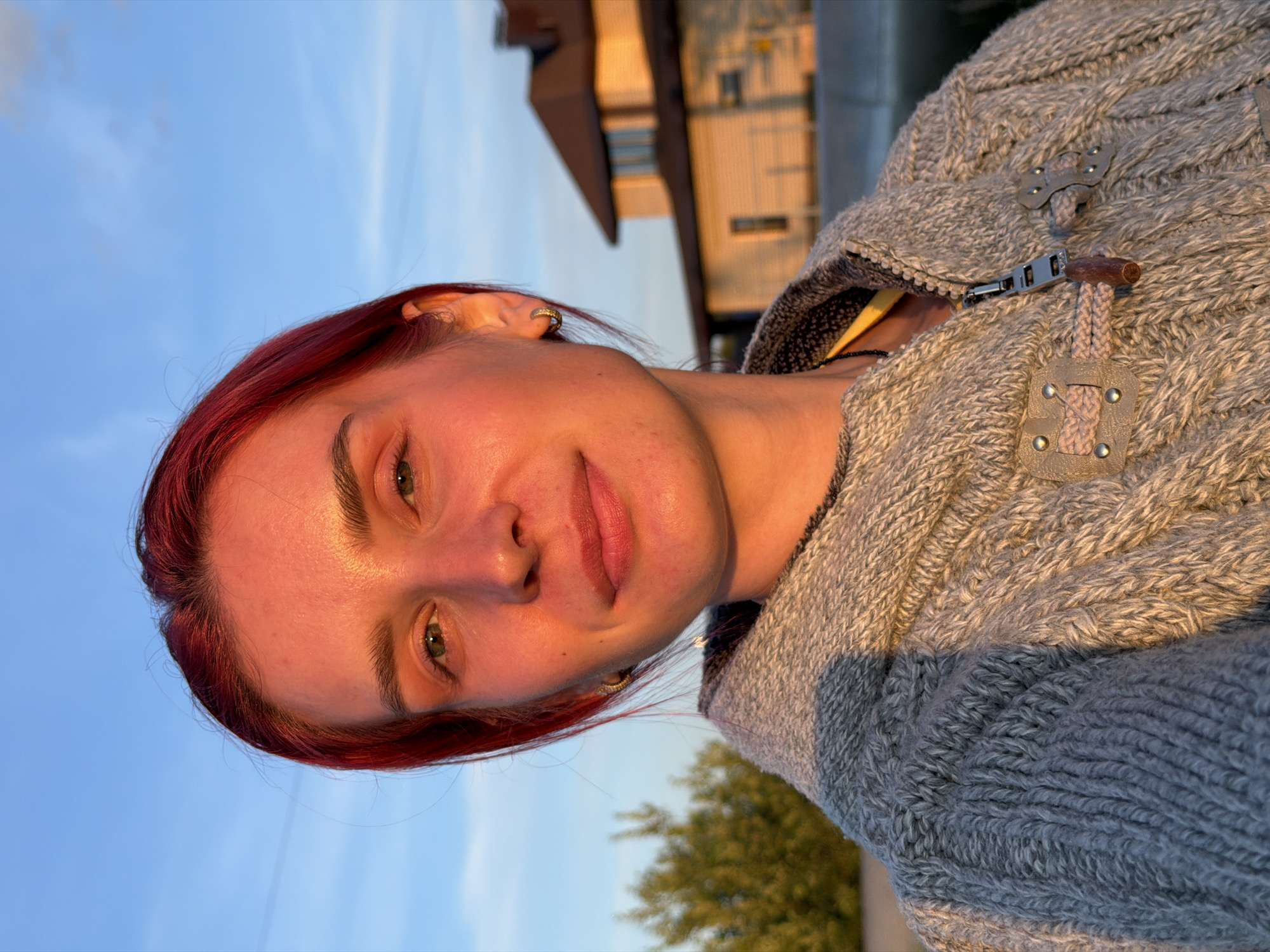 A young woman with bright red hair, wearing a gray knitted jacket, is captured in a close-up selfie outdoors. The setting appears to be suburban, with a house visible in the background and trees to the left. The warm, golden light suggests it's either sunrise or sunset. The sky is a clear blue with a few wispy clouds. The woman has green eyes and is wearing small hoop earrings. The image is well-lit, with a focus on the woman's face and upper body. There are no discernible elements that specifically place the location in Chaikovsky, Russia, beyond the user's prompt.