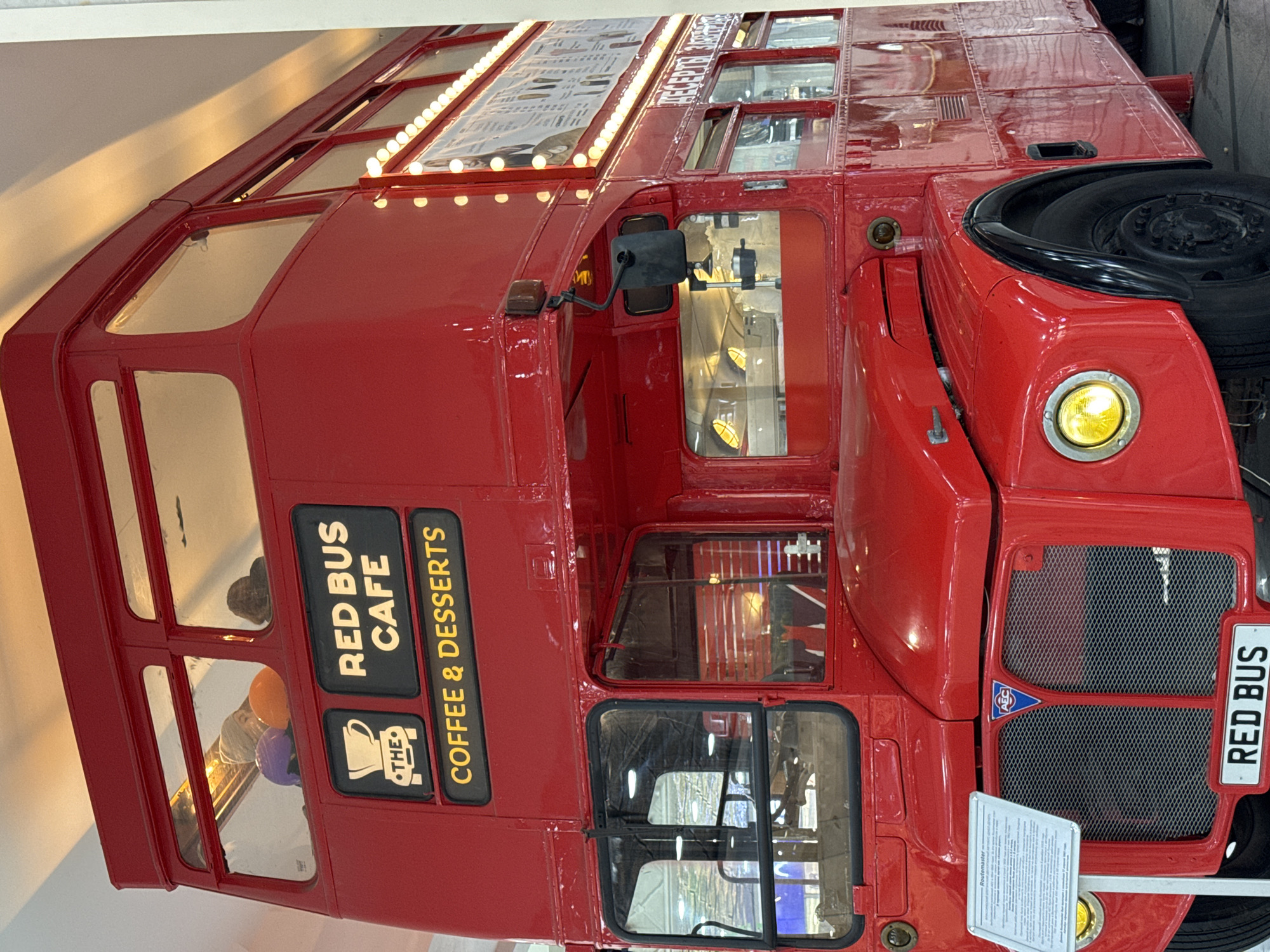 A red double-decker bus, labeled "RED BUS CAFE" and "COFFEE & DESSERTS" is on display in what appears to be a museum or exhibition space. The bus has windows on both levels, a black side mirror, and a black spare tire mounted on the front. The front grill of the bus has a "RED BUS" license plate and a sign with text. The bus is in Yekaterinburg, Russia.