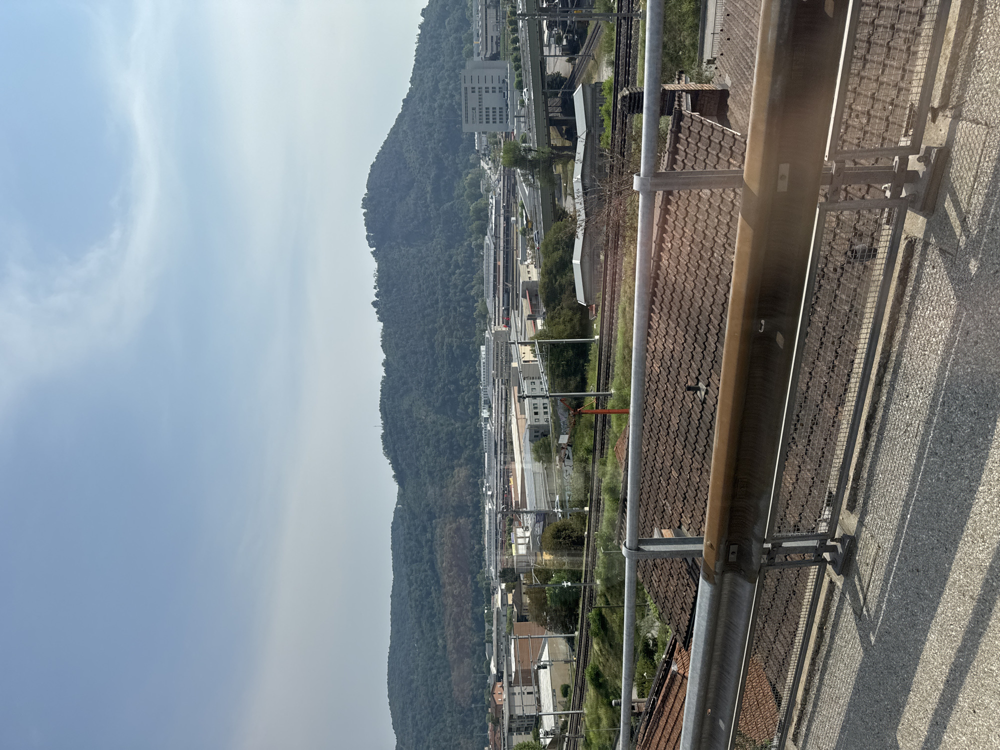 The image depicts a panoramic view of an industrial area and a forested hill under a clear, lightly clouded sky. In the foreground, a metal guardrail with a rusty section runs horizontally across the frame, casting shadows on a textured paved surface. Below the guardrail, red-tiled rooftops of buildings are visible.
The middle ground features a complex of industrial buildings and infrastructure. A set of railway tracks runs from the left towards the center. Several low-rise industrial structures with light-colored facades and dark roofs are scattered throughout the area. A multi-lane overpass with cars on it spans across the railway lines and industrial buildings.
In the background, a large, densely forested hill dominates the landscape. The hill appears to be a significant natural feature within or bordering the urban environment. The sky above is a pale blue, with wispy white clouds. The scene is brightly lit, suggesting it was taken during daylight hours. The overall impression is one of an industrial zone situated at the edge of a natural, hilly terrain.