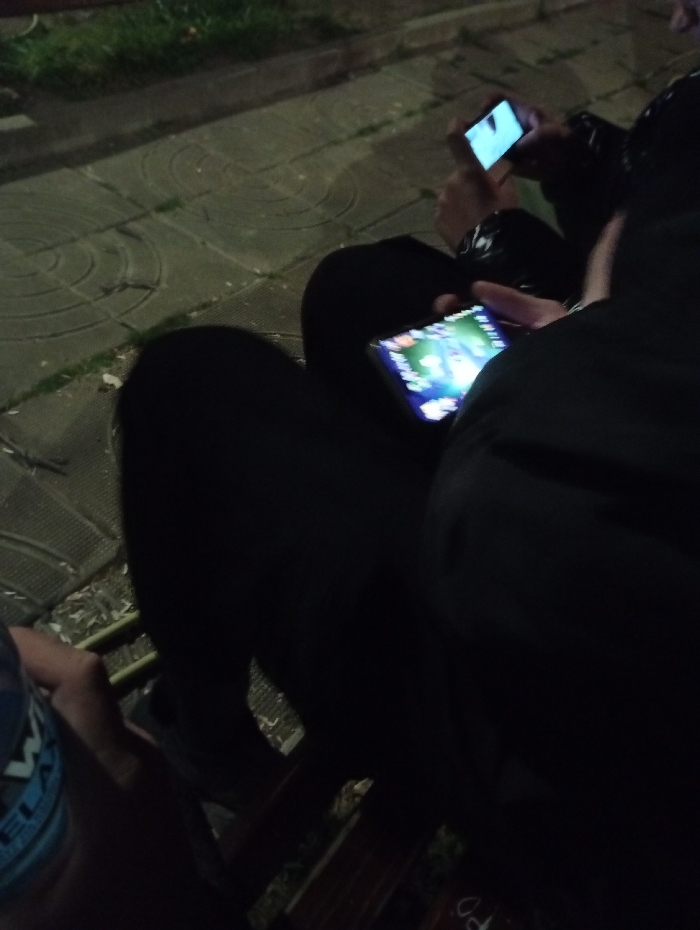 The image depicts at least three individuals seated outdoors at night or in a dimly lit area. The ground surface consists of concrete paving stones with visible circular patterns, adjacent to an area of grass. In the foreground, the left arm and a portion of a blue and white patterned shirt of one person are visible. Adjacent to this individual, another person is seated, wearing dark clothing, and holding a smartphone with both hands. The smartphone screen is illuminated, displaying a colorful graphical interface, consistent with a mobile game. Further back, a third individual is partially visible, also wearing dark clothing, and holding a smartphone with both hands, its screen similarly illuminated with a bright display. A wooden bench or seating structure is partially visible in the lower section of the image, featuring a white marking. All visible individuals are engaged with their respective mobile devices. The specific location within the city of Lebnitsa, Bulgaria is not discernible from the visual information provided.