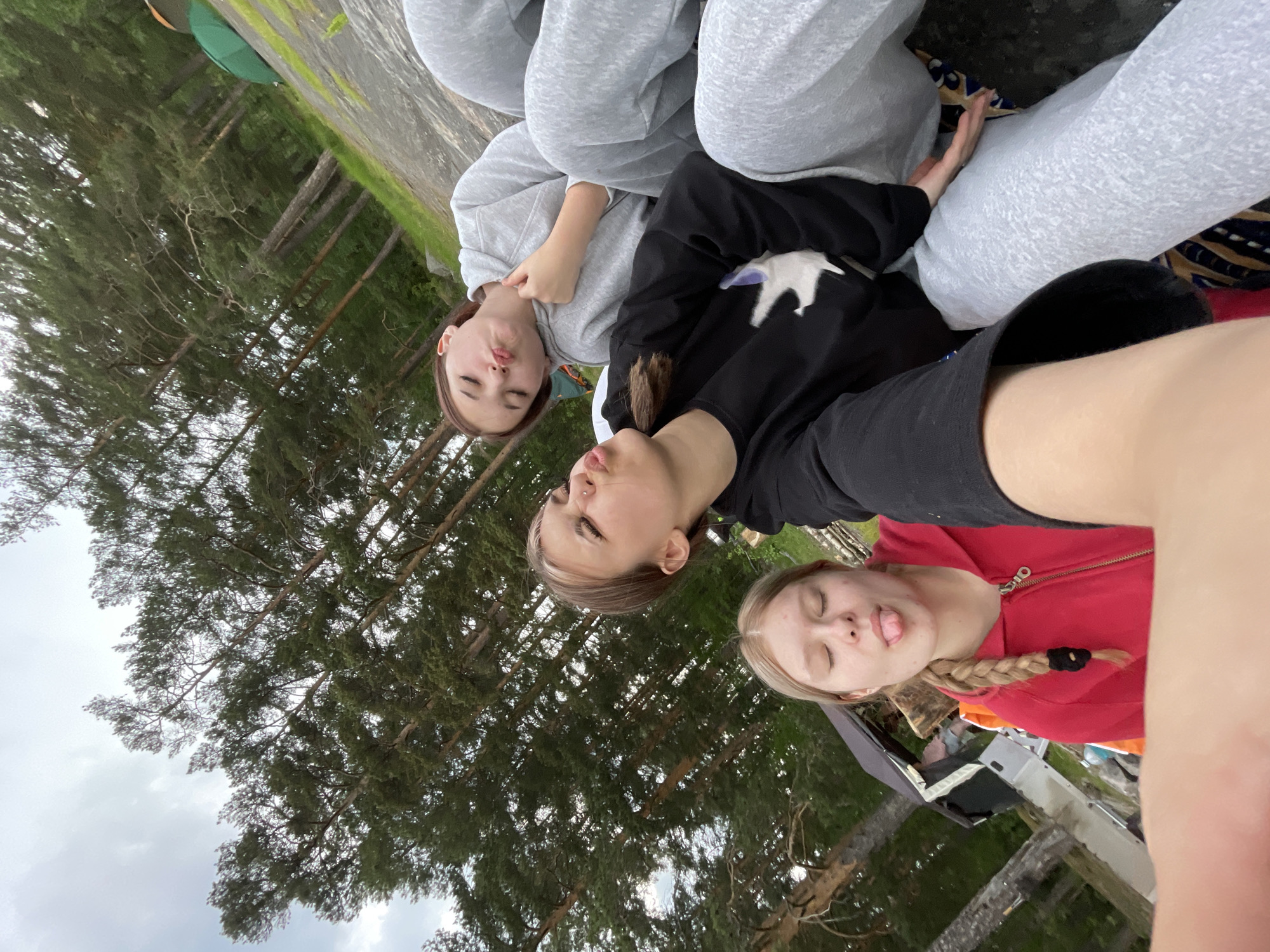 This image captures a selfie featuring three young females in an outdoor, natural environment near Boitsovo, Russia. The camera is held low, providing an upward-angled view of the subjects and the surrounding landscape.
In the foreground, centered on the left, a female with blonde hair styled in a braid to her right side is visible. She is wearing a red zip-up hooded jacket, has her eyes closed, and her tongue extended playfully. Her right arm, likely holding the camera, is partially visible at the bottom of the frame.
To her right and slightly behind, another female is positioned. She has light brown hair with lighter streaks and a nose piercing. She wears a black long-sleeved top featuring a white graphic design. Her eyes are partially closed, and her lips are pursed in a "fish face" expression.
Further to the right and slightly behind the second individual, the third female is visible. She has darker hair and is dressed in a light grey hooded sweatshirt and matching light grey sweatpants. Her eyes are also mostly closed, and her lips are pursed in a similar playful "fish face" expression. Her right hand is positioned near the collar of her hoodie.
The three individuals are in close proximity, suggesting camaraderie. They are seated or reclining on what appears to be a patterned blanket or fabric laid over rocky ground.
The background is dominated by numerous tall pine trees with dark green foliage and reddish-brown trunks, indicating a dense forest or woodland. The sky above is overcast, showing light grey clouds. The ground is uneven, composed of rocks with patches of green grass and moss. In the far background, partially obscured by trees and terrain, two tents are visible, one predominantly green and another with a brownish hue, suggesting a camping or outdoor recreational setting within the forest.