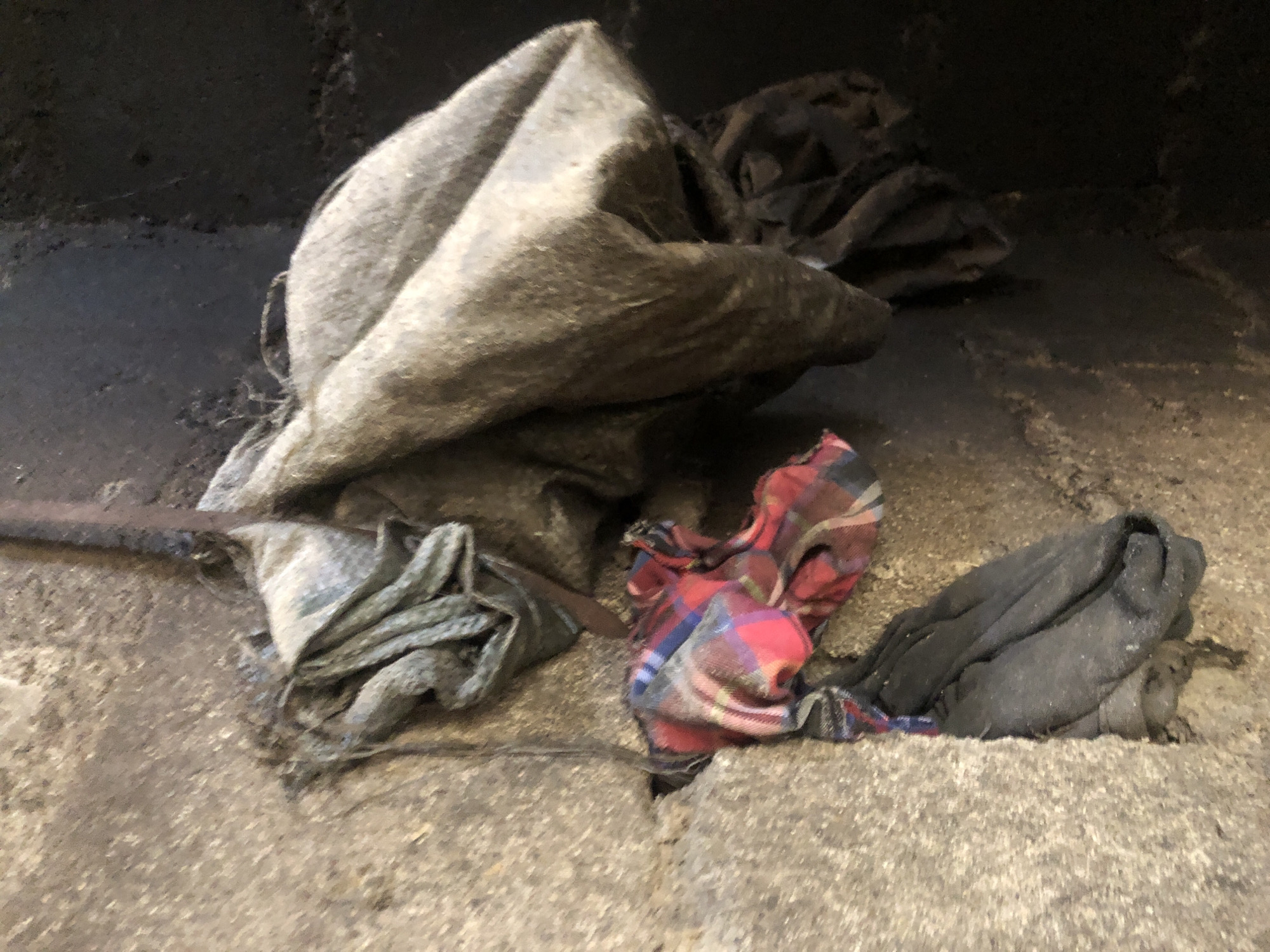 The image shows a collection of dirty rags and fabrics piled on a rough, gray concrete surface. The scene appears to be indoors, possibly in a workshop or storage area, with a dark, textured background that suggests a wall or enclosure. The lighting is dim, creating shadows and highlighting the textures of the materials.
The main subjects are several pieces of fabric. The largest is a gray, coarse-textured rag, folded and crumpled, occupying the central and upper left portion of the image. To its right, a piece of red, blue, and black plaid fabric is folded, revealing its checkered pattern. To the far right, a dark gray or black rag is similarly piled. In the lower left, another gray rag with frayed edges is visible, attached to a thin, dark rod or handle. The rags appear to be well-used and dirty, suggesting they have been used for cleaning or some other practical purpose.
The arrangement of the rags is haphazard, giving the impression of a pile of discarded or stored items. There is no clear activity taking place; the image captures a still moment of these objects in their setting. The overall mood is utilitarian and somewhat grimy due to the state of the fabrics and the rough environment. No text is visible in the image. The time of day and weather are not discernible from the scene.