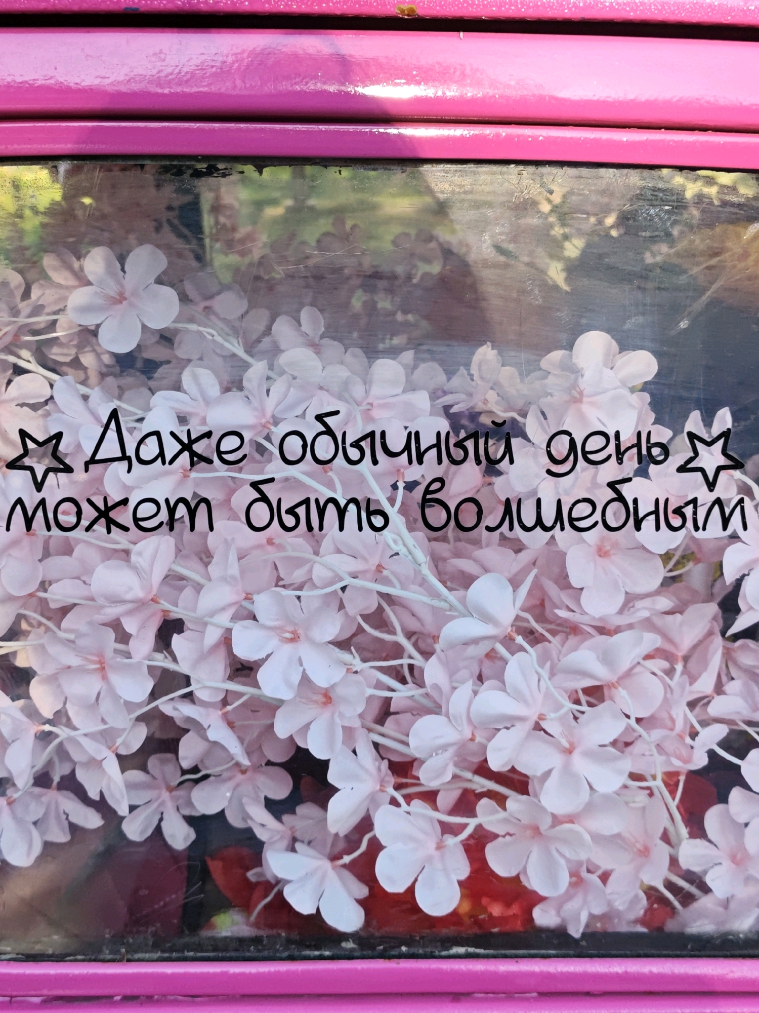 The image shows a close-up of a pink, metallic surface with horizontal indentations. Below this surface, a clear pane of glass reveals an arrangement of artificial pink cherry blossoms. Black handwritten text in Russian is overlaid on the flowers. The text reads: "Даже обычный день может быть волшебным" which translates to "Even an ordinary day can be magical." The background behind the flowers is blurred and appears to be outdoors with hints of green foliage and possibly red objects. The overall impression is a decorative display. There is no discernible information in the image that would suggest a specific location in Moscow, Russia, or any people or actions taking place.