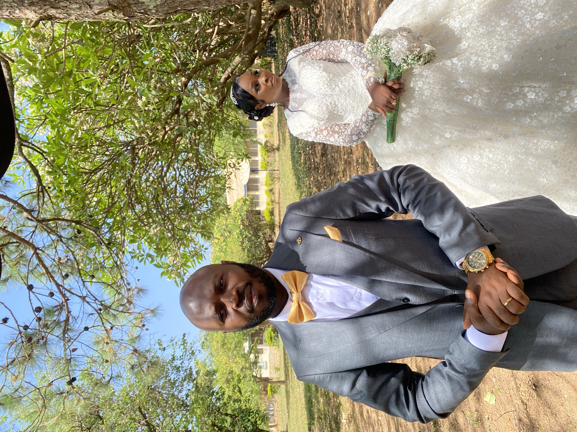A full-length, slightly rotated, outdoor shot features a couple in wedding attire, likely in Dutsen Kato, Nigeria. The groom is in the foreground, wearing a gray suit with a gold bow tie and pocket square. He has a gold watch on his left wrist and is clasping his hands in front of him. The bride is positioned behind and to the right of the groom. She wears a full-length white gown and holds a bouquet of white flowers. The background includes trees, grass, and a building in the distance. The scene is bathed in natural daylight.