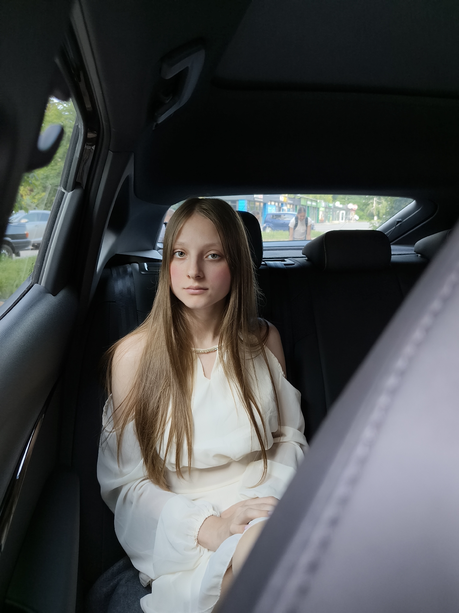 A young female individual is seated in the rear left passenger seat of a vehicle, looking directly at the viewer. She has long, straight, light brown hair, fair skin, and light-colored eyes. Her cheeks display a subtle reddish tone, and she wears a thin, gold-toned necklace. She is dressed in an off-white, off-the-shoulder garment with long, loose sleeves, and her hands are clasped in her lap.
The vehicle's interior is dark, with black or dark grey upholstery on the seats, headliner, and door panels. Grab handles are visible on the ceiling, and a seatbelt is present on the unoccupied seat to the individual's right. The image is captured from a perspective within the front of the car, looking towards the rear.
Through the rear window, an urban street environment identified as the city of Kryvyi Rih, Ukraine, is visible under daylight conditions. The background shows a commercial street lined with multi-story buildings, some featuring blue and green architectural elements on their storefronts. Several vehicles, including a blue car and a lighter-colored sedan, are present on the street. Trees with green foliage are visible along the street and in the distance. A pedestrian, wearing a light-colored shirt and a backpack, is observed walking on the sidewalk in the mid-ground. No direct interactions are depicted between the seated individual and the exterior environment or other individuals.