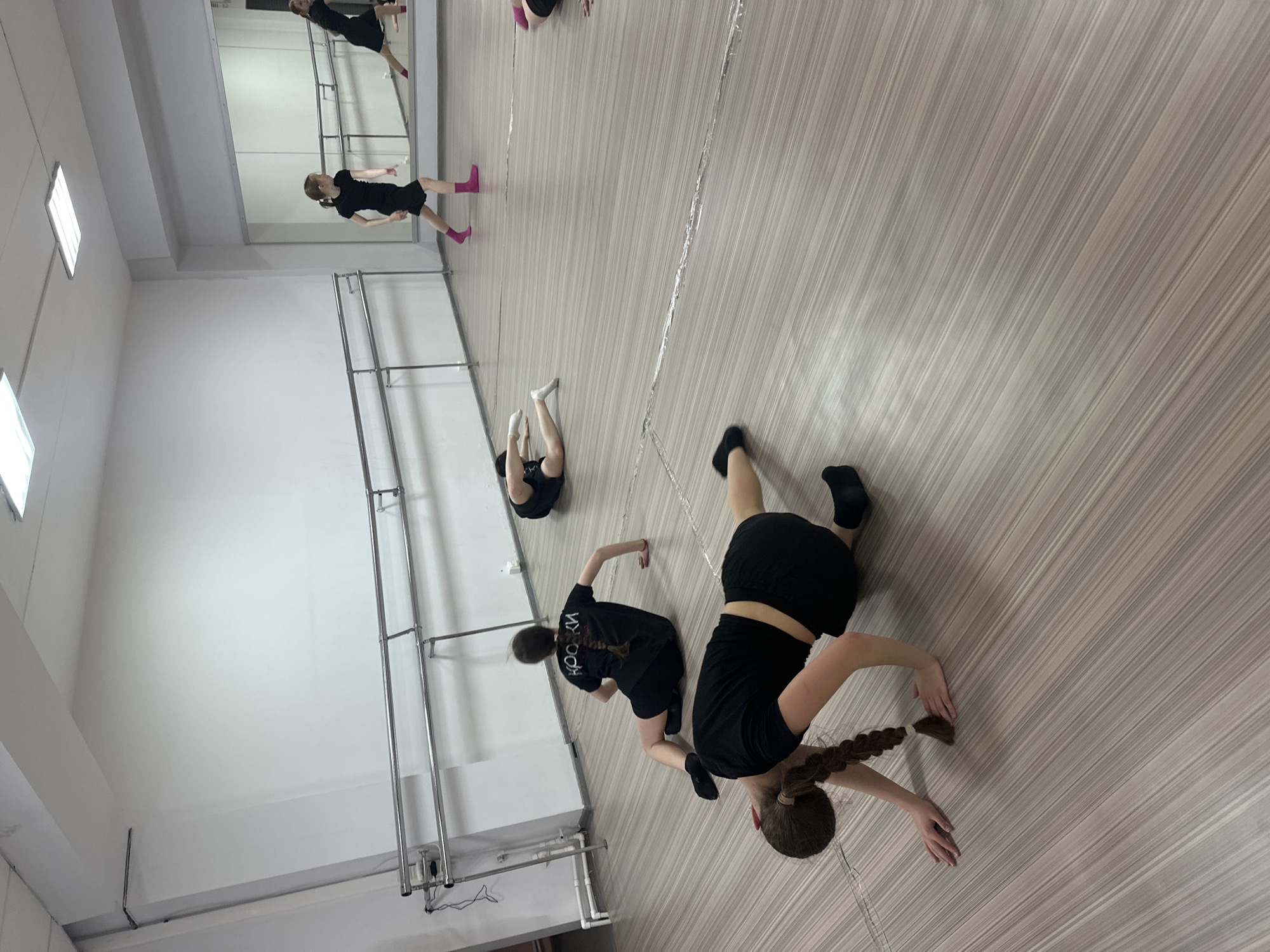 The image shows a dance studio in Votkinsk, Russia. Five individuals are present, engaged in stretching exercises on a light wood-grain floor. The studio has white walls, a ballet barre, and a mirrored wall reflecting the dancers. Four of the dancers are wearing black clothing, while one wears black with white socks. A section of metallic-looking ribbon or tape runs along the floor.