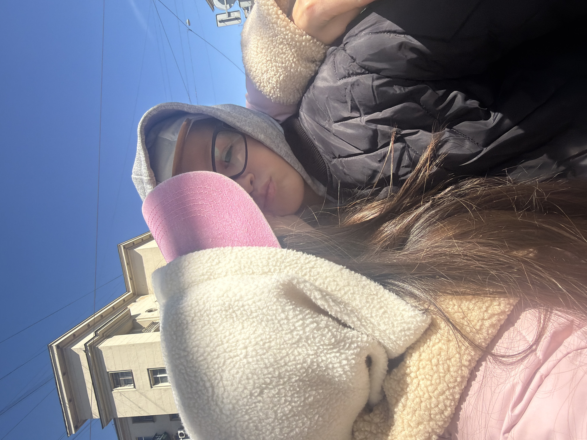 A close-up outdoor portrait features a person, likely a young woman, looking slightly upwards towards the camera against a clear blue sky. She wears a grey hooded sweatshirt, black-rimmed glasses, and a vibrant pink baseball cap. Over the sweatshirt, she has on a black puffy jacket with a cream-colored, fluffy sherpa lining visible around the collar and hood. Her long, dark brown hair falls over her shoulders. The person's expression is calm and neutral, with a slight pout.
The bright sunlight suggests a clear day, likely morning or afternoon. In the background, to the person's left, a light-colored building with classic architectural elements, including cornices and windows, stands against the sky. This architectural style is characteristic of Saint Petersburg, Russia, aligning with the provided location context. Thin utility lines are also visible stretching across the blue sky. There is no visible text in the image.