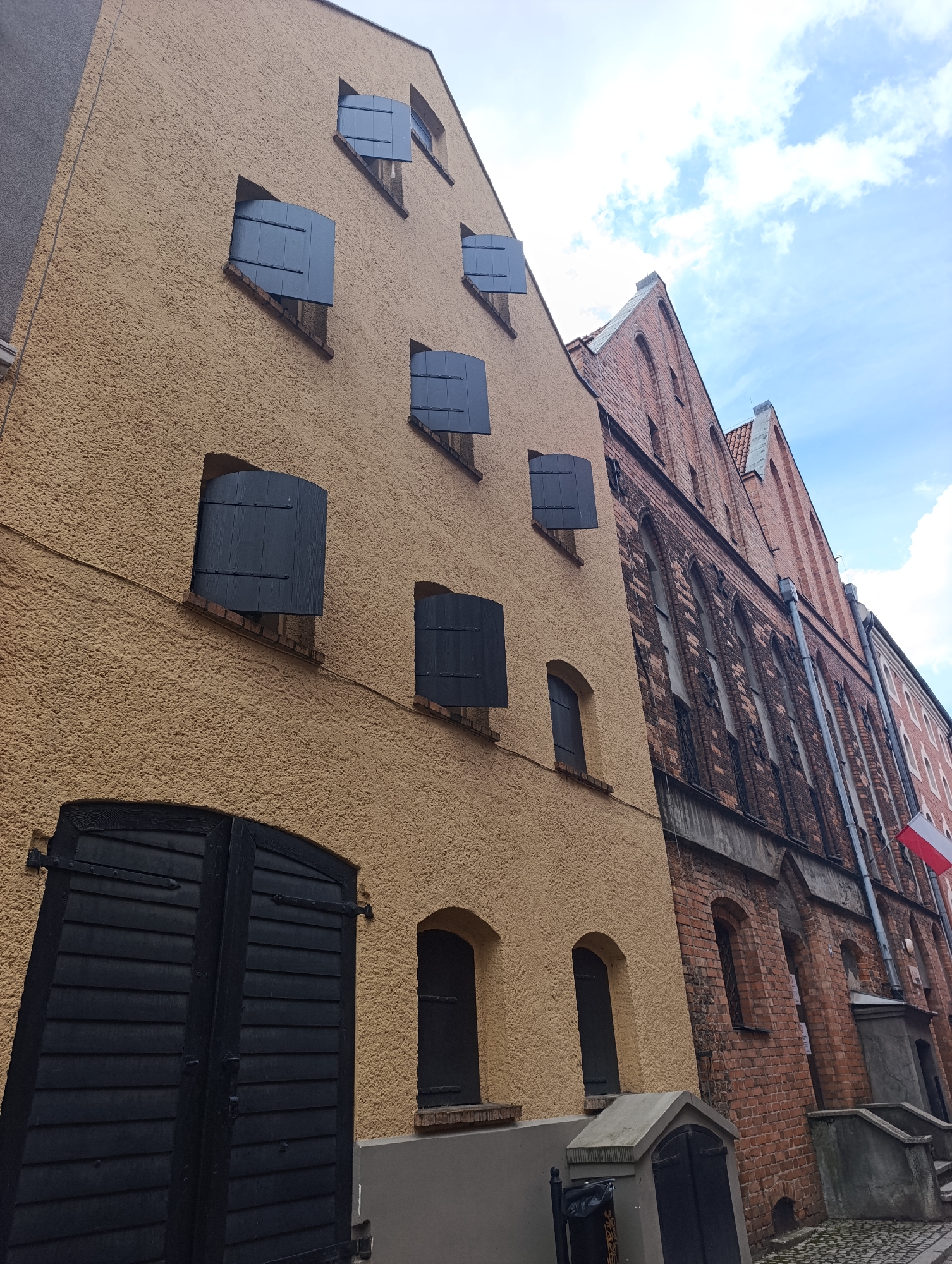 The image shows a street view in Toruń, Poland, featuring historic buildings with distinctive architecture. On the left, a building with a textured ochre facade has multiple windows with dark shutters, some partially open, and a large set of black wooden doors at street level. To the right, a taller, older building constructed of red brick displays a series of arched windows with dark frames and a steeply pitched gabled roof. The street is paved with cobblestones. The sky above is partly cloudy with a bright blue hue. The architectural style suggests a historical district, likely within the Old Town of Toruń.