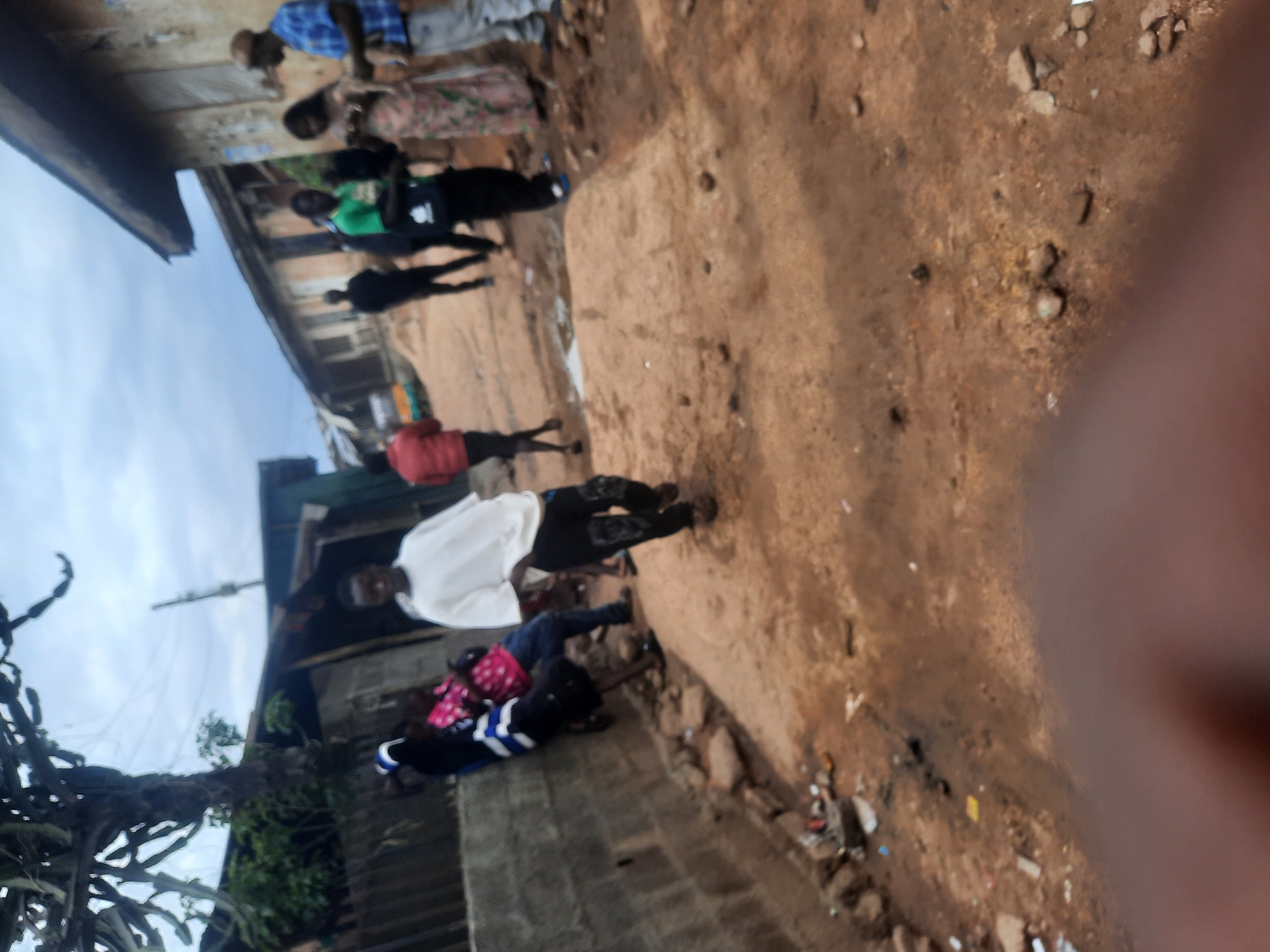 This is a blurry, slightly rotated outdoor image depicting a street scene in Gwong, Nigeria, under an overcast sky. The primary subjects are approximately seven to nine individuals scattered along an unpaved, dirt path.
In the foreground, to the right, a blurry brown thumb or finger partially obscures the view of the rough, reddish-brown ground, which is littered with small rocks and various pieces of debris, including what appears to be plastic and paper.
In the midground, several people are present. A man in a white long-sleeved shirt and dark pants stands upright, looking slightly over his shoulder. Near him, two figures are lower to the ground, possibly sitting or crouching; one wears a dark top with pink patterned details, and the other has a dark top with white and blue stripes on the sleeves. Further back, another person in a red top is visible. Beyond them, a person wearing a bright green shirt and carrying a black bag walks past. In the background, under a building, a man in a blue plaid shirt and light pants stands near a woman with long dark hair dressed in a patterned garment. Other blurry figures are partially visible in this group.
The setting appears to be a residential or semi-urban area. Old, two-story buildings with faded paint line the dirt path, suggesting dwellings or small businesses. To the far left, a sparse, gnarled plant, possibly a type of cactus or old tree, stands prominently against the cloudy sky. The overall activity seems to be casual, with people either standing, walking, or resting along the path.
The time of day is daytime, indicated by the light, but the visible overcast sky suggests either an early morning, late afternoon, or simply a cloudy day. Due to the blurriness of the image, no specific emotions are discernible, and no visible text can be read.