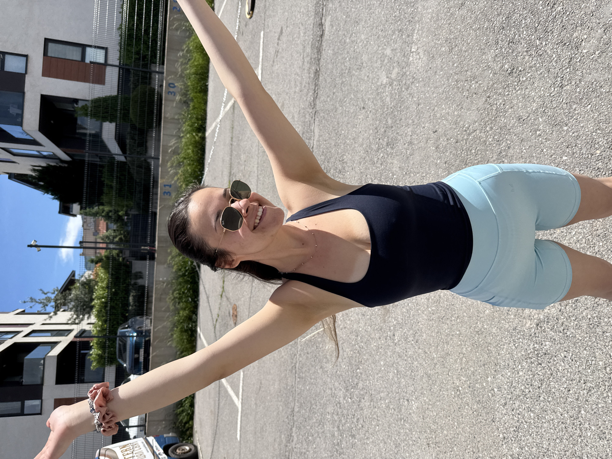A woman in a black tank top and light blue athletic shorts is standing in an outdoor parking lot, smiling and holding her arms outstretched. She is wearing sunglasses and a bracelet. The parking lot has painted white lines dividing parking spaces, and a dark gray asphalt surface. In the background, there is a modern residential building with dark windows and a fence along the property line, with vegetation behind the fence. The image has rotated during processing, making it appear sideways. The location is likely in Sofia, Bulgaria.