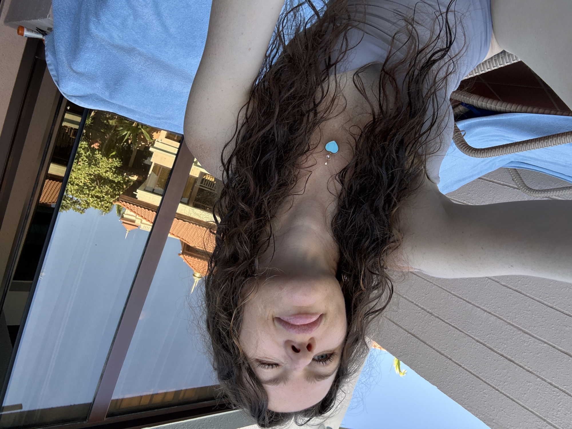 A woman with long, wet, curly brown hair and fair skin is captured in a close-up selfie, seated outdoors in a woven lounge chair. She is wearing a light-colored swimsuit top and a gold-toned necklace featuring a turquoise heart-shaped pendant with a small clear gemstone. Her right eye is winking, while her left eye is open, and her lips are slightly parted. The lounge chair is partially covered by a light blue terry cloth towel. Behind her, a large, dark-framed window reflects a multi-story building complex. The reflection displays structures with light-colored facades, terracotta-tiled roofs, balconies, and distinctive ornate finials. Palm trees are also visible in the reflection. A white and orange bottle is situated on a ledge to the right of the blue towel. The architectural characteristics visible in the reflection strongly suggest the Jumeirah Al Qasr hotel within the Madinat Jumeirah complex in Dubai, United Arab Emirates. The scene is illuminated by bright sunlight under a clear blue sky.