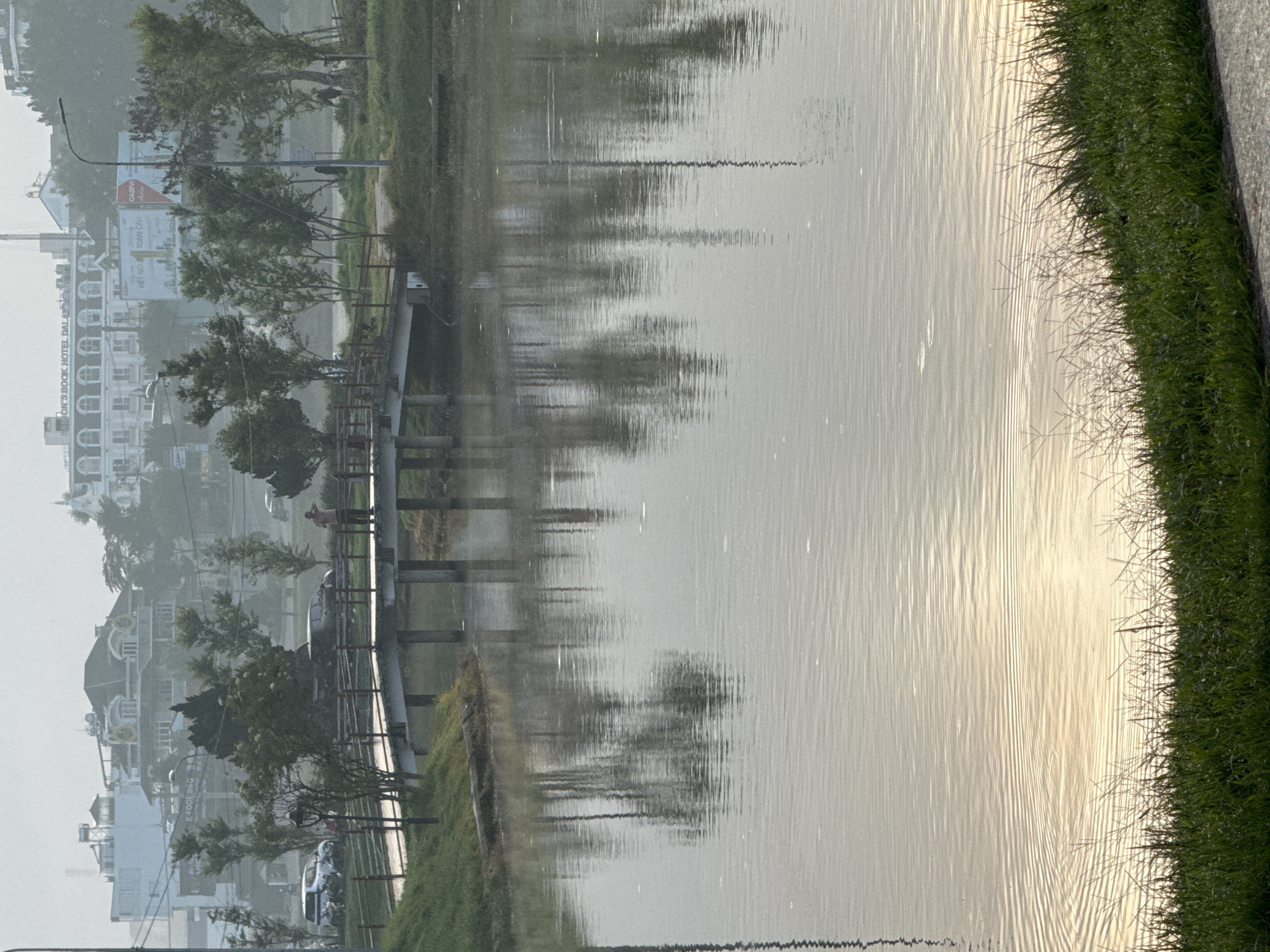 The image shows a tranquil view of Xuân Hương Lake in Đà Lạt, Vietnam, on a hazy morning. A pedestrian bridge crosses the water, with two people standing on it. The reflections of trees and buildings are visible on the lake's surface. The foreground features lush green grass and a paved path. In the background, a row of trees separates the lake from a cityscape featuring several buildings, including a prominent hotel. The overall atmosphere is calm and serene, with a soft, diffused light indicating early morning or late afternoon.