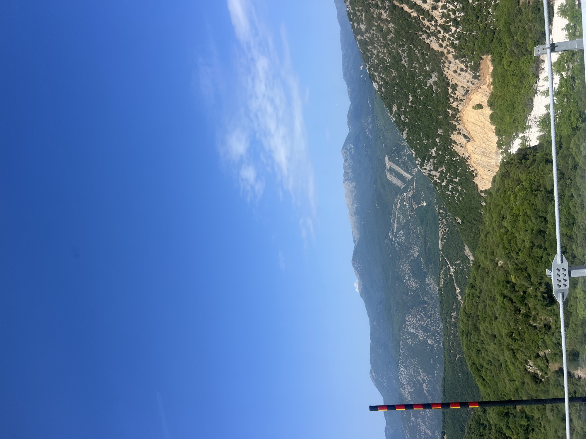 The image captures a vast mountain landscape under a clear blue sky with scattered white clouds. In the foreground, a black pole with alternating red and yellow bands is visible on the left side, alongside a wire fence. The mountains are covered in lush green vegetation, with some areas showing exposed rock and soil, particularly on the right side where a landslide appears to have occurred. The distant peaks are tinged with a lighter shade, suggesting haze or distance. The scene is indicative of a natural, undeveloped area, possibly a mountain road or viewpoint in the vicinity of Νέος Γοργόμυλος, Greece, given the context. There are no people or animals visible in the image.