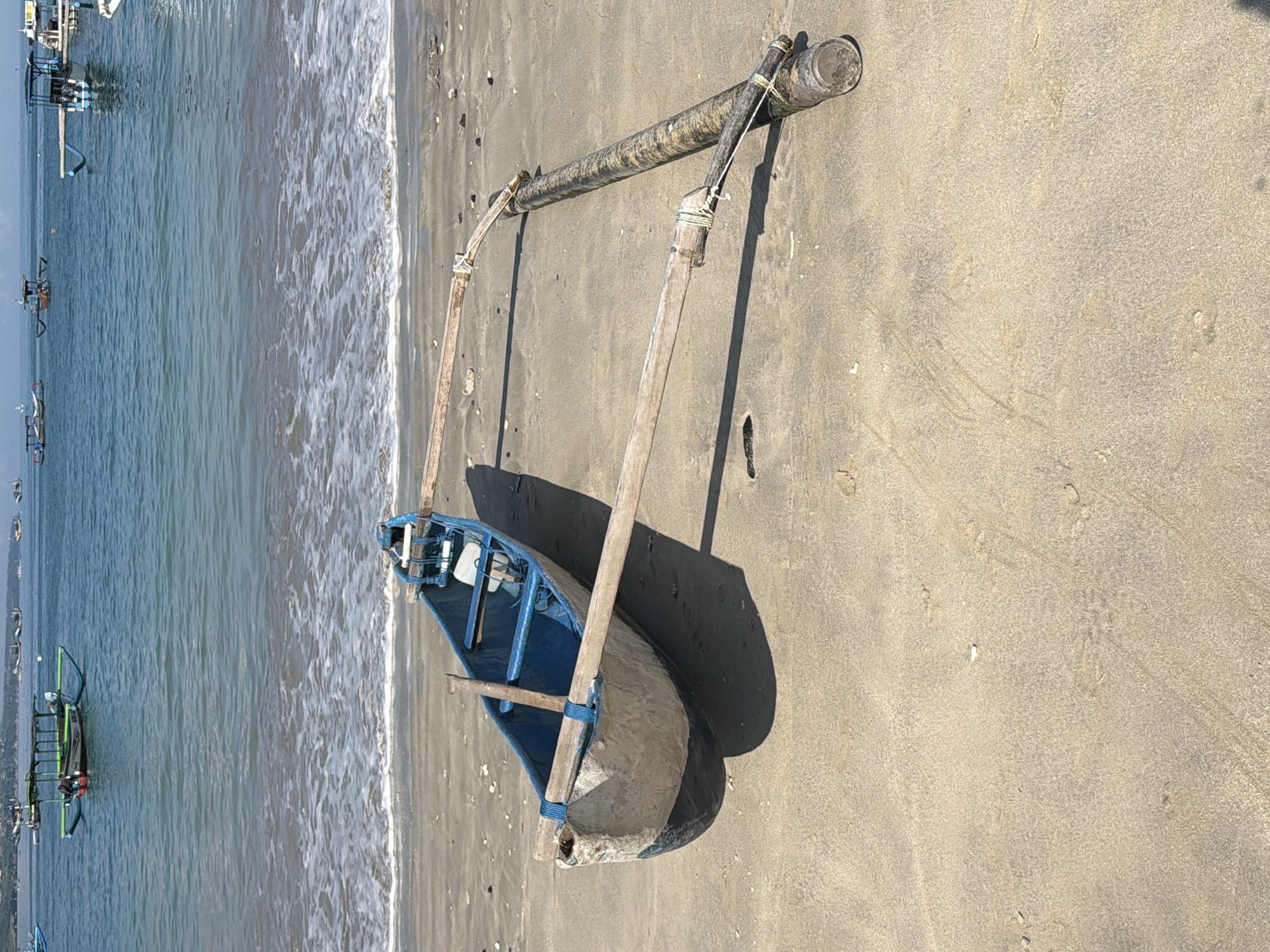 The image shows a traditional Indonesian outrigger canoe, specifically a Jukung, resting on a sandy beach. The canoe is blue and has two wooden outriggers connected to a long, cylindrical float with rope. The beach is light tan, likely sandy, and has some darker patches. The ocean is visible in the background, with gentle waves lapping the shore and other boats docked in the background.
Based on the presence of Jukungs, a hallmark of Balinese coastal life, and the overall beach environment, the location is likely Kuta, Indonesia, or a nearby coastal area in Bali. The scene likely depicts a typical fishing boat or tourist transport vessel temporarily beached.