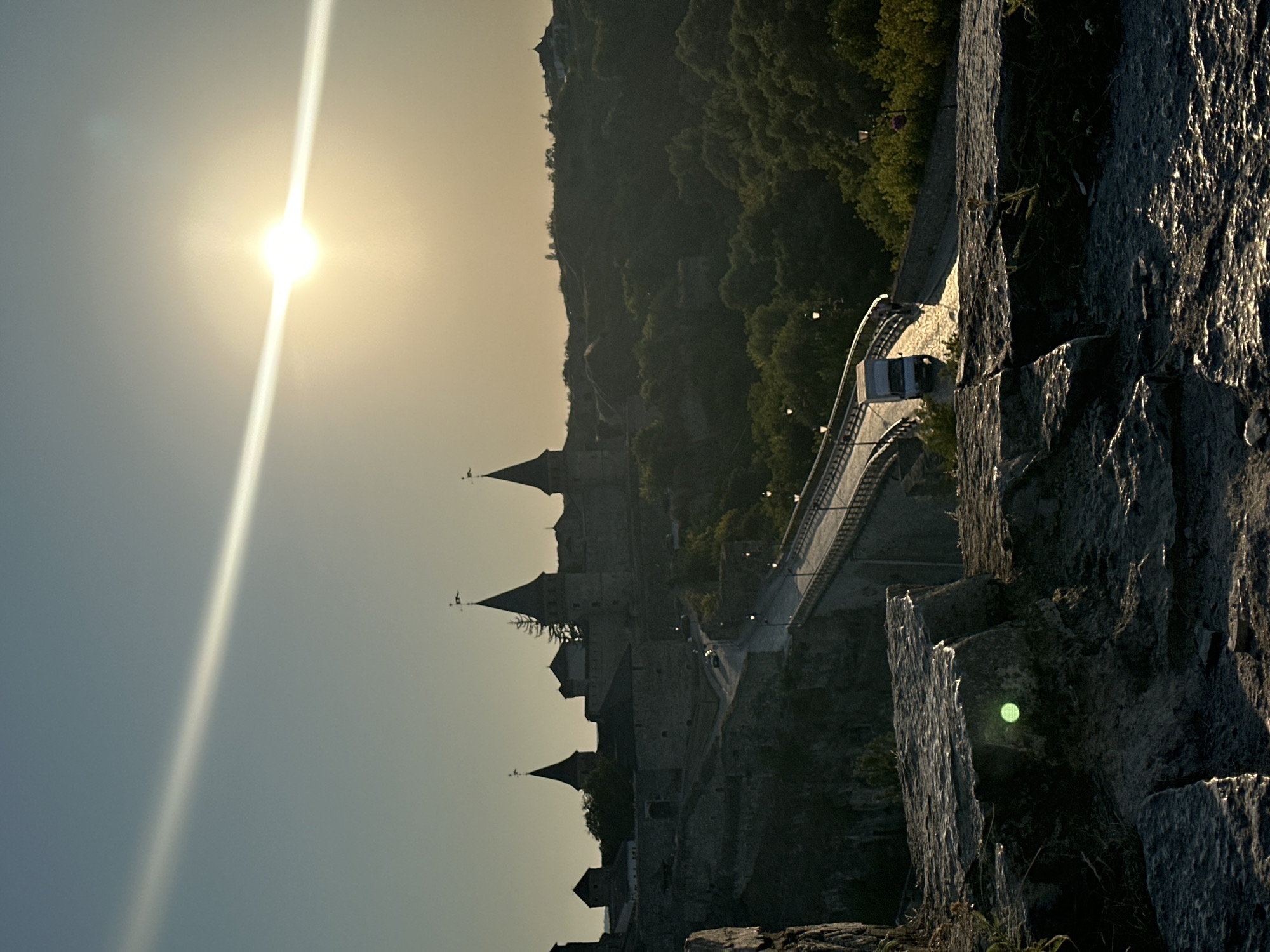 The image captures a scene in Kamianets-Podilskyi, Ukraine, featuring the Kamianets-Podilskyi Castle and the adjacent New Polish Bridge at sunset. The sun is low in the sky, creating a bright glare that streaks across the upper portion of the image, with visible lens flare. The castle, a prominent stone structure with multiple conical towers, is silhouetted against the fading light and set upon a verdant, tree-covered hillside. The New Polish Bridge, a modern structure with a curved roadway, spans a gorge and is illuminated by streetlights. A white van is visible driving across the bridge. In the foreground, large, rough-hewn stones are visible, with a greenish light reflection on one of them. The overall impression is one of historical grandeur bathed in the dramatic light of sunset.