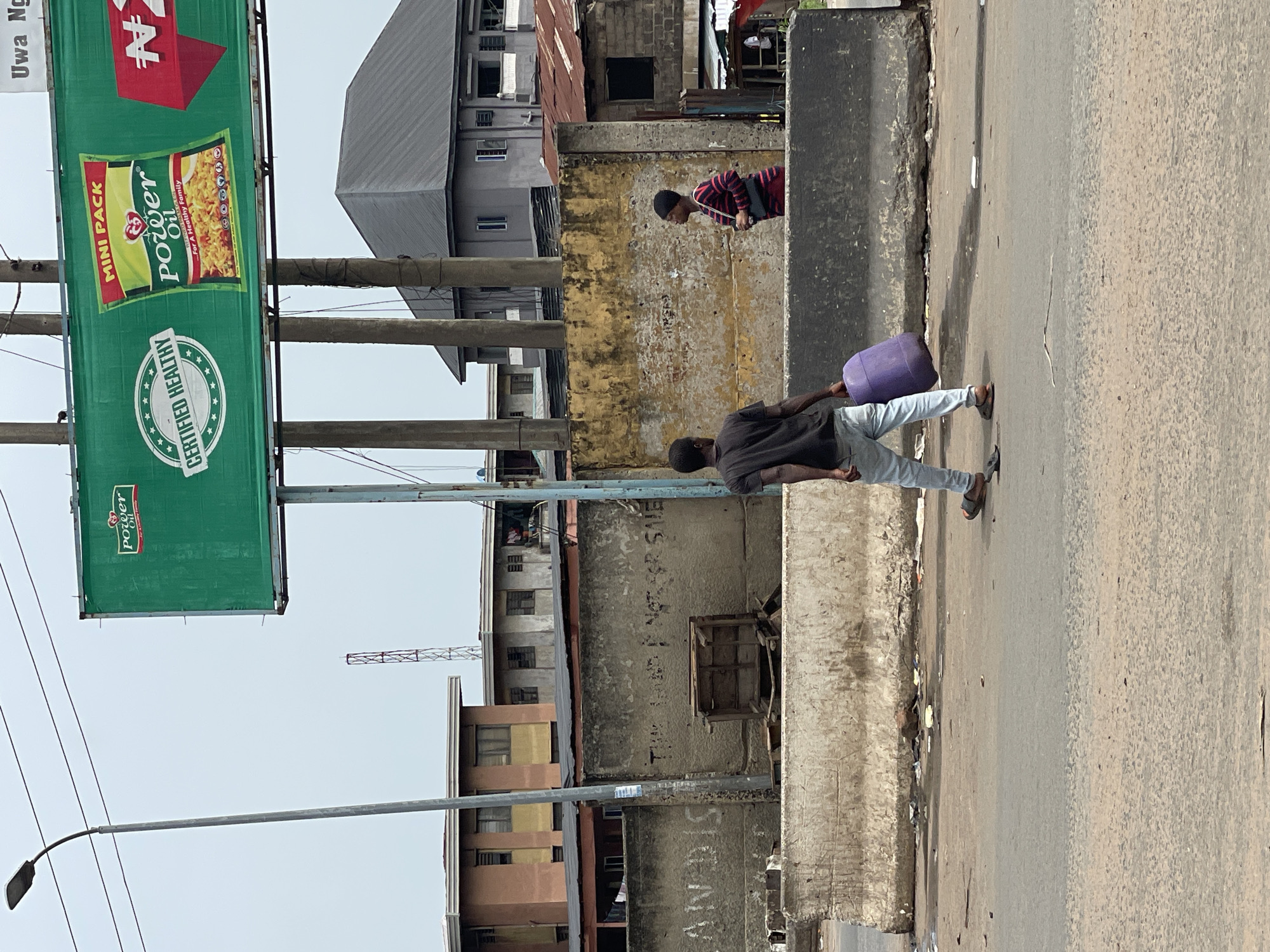 The image depicts a street scene in Onitsha, Nigeria. An adult male is walking along the edge of the road carrying a purple jerrycan. A second person is visible on the opposite side of a concrete barrier. In the background, there is a mix of residential and commercial buildings, some appearing dilapidated. An advertisement for Power Oil is prominently displayed on a green billboard, with the words "CERTIFIED HEALTHY" and "MINI PACK" visible. The street appears dusty and littered. A streetlight is visible in the background. The general atmosphere suggests a low-income urban environment.