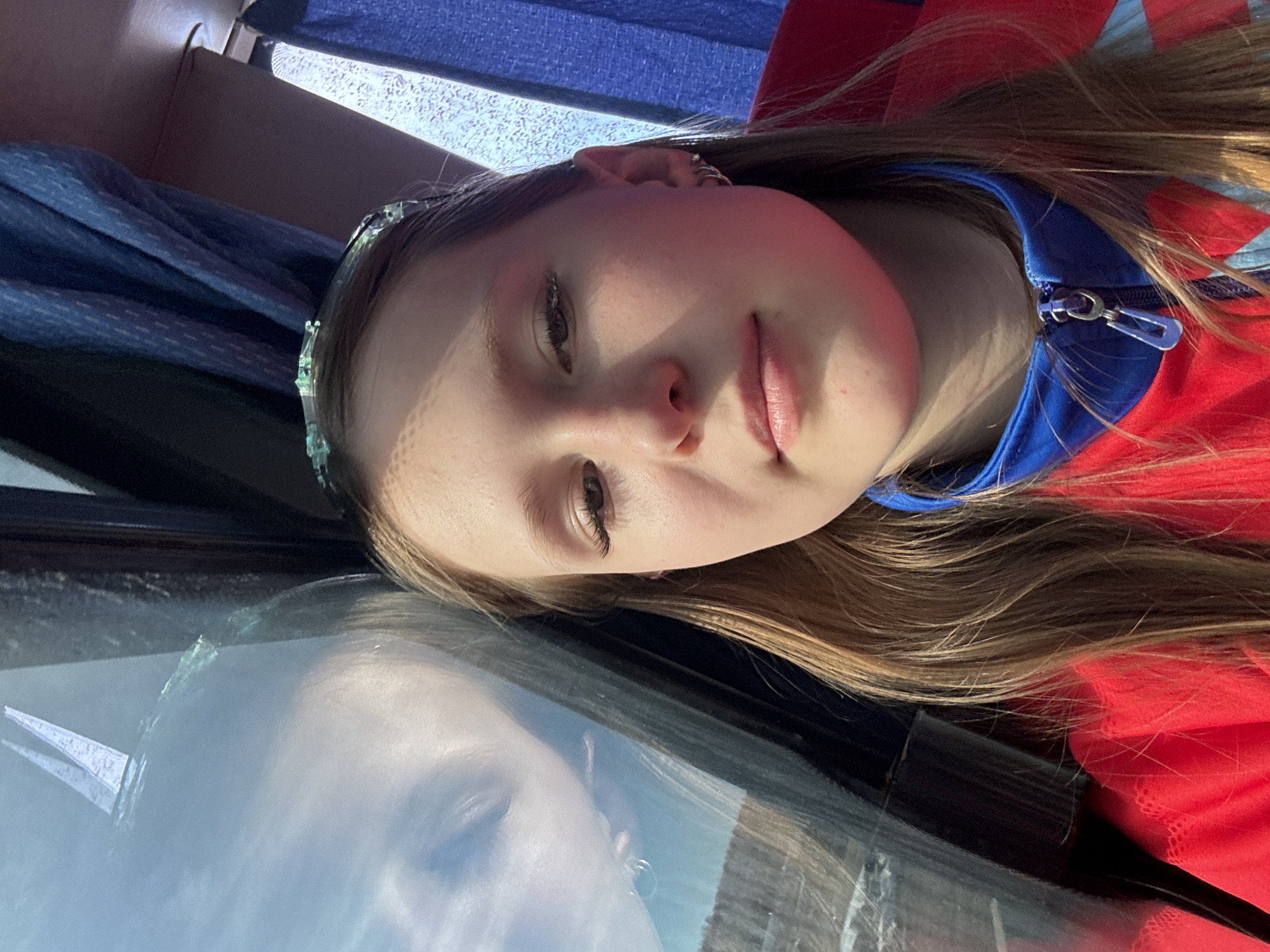 A close-up shot depicts a young girl with light skin and brown hair. She is wearing a red and blue zip-up jacket and a decorative headband. Her face is partially reflected in a window. The backdrop shows a blue curtain, suggesting she is inside a vehicle, potentially a bus or car. No specific details visible indicate that this scene is located in Adishchevo, Russia.