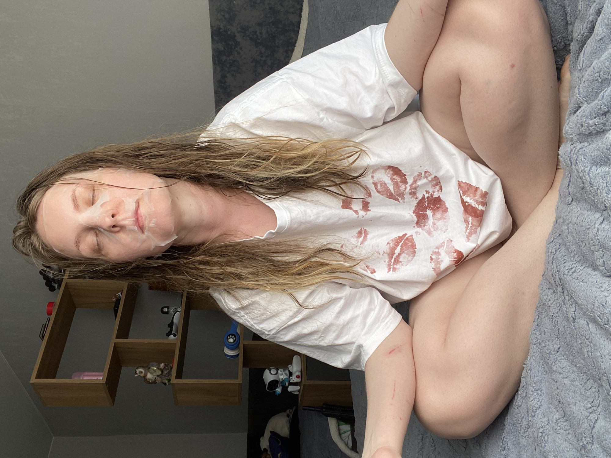 A person is sitting cross-legged on a bed covered with a grey blanket. The person is wearing a white t-shirt with multiple red lipstick kiss prints on the front. The person has fair skin, long blonde hair and is wearing a facial mask. There are visible scratches on both arms. In the background is a wall with a wooden shelf unit holding various small objects. The location is likely inside a residential dwelling. There is no verifiable location in Noviy, Russia.