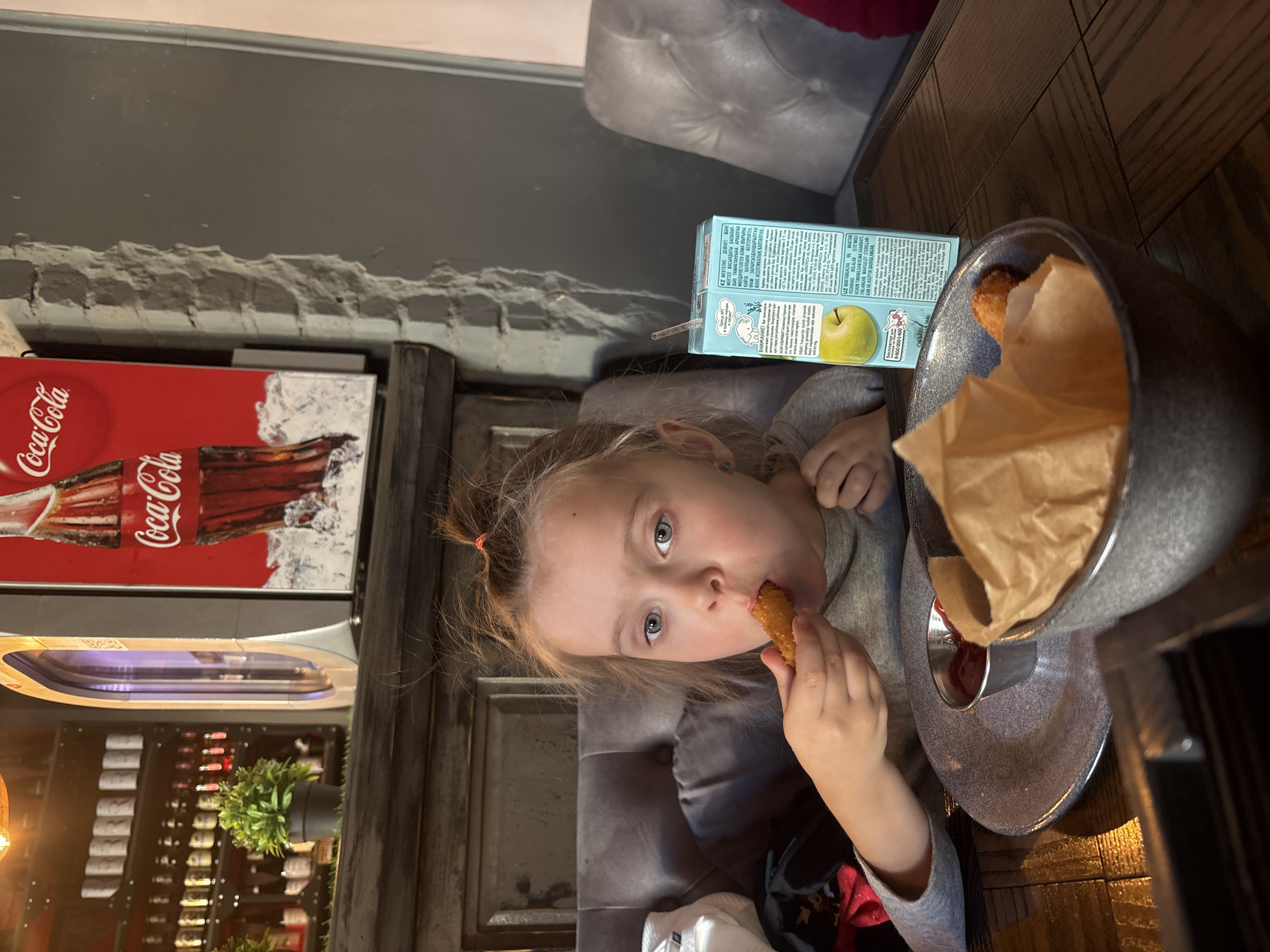 The image is an overhead shot of a young girl eating at a restaurant. The girl, with light skin and blue eyes, is seated on a gray cushioned bench. She is wearing a gray long-sleeved shirt and has her hair pulled back. She is holding a breaded nugget in her right hand and has it near her mouth.
On the wooden table to her right, there's a blue juice box with an apple on it, a gray bowl containing paper-lined fried food, and a small dish of ketchup.
In the background, there is a Coca-Cola sign and a display of bottled wine. The lighting in the room appears dim. The location is the city of Perm, Russia.