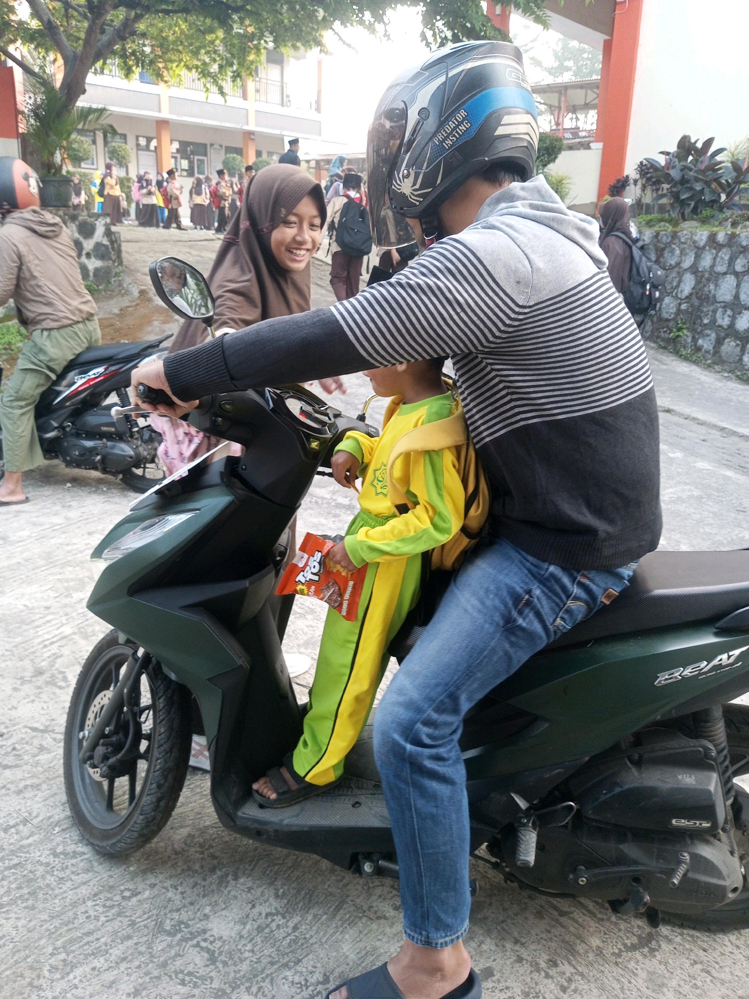The image shows two people, a child and an adult, on a dark green scooter in an outdoor setting that appears to be a school yard or a public area. The child, wearing a bright yellow and green uniform with a yellow backpack, is seated in front of the adult. The adult, wearing a grey and black striped hoodie and blue jeans, is driving the scooter and wearing a black and blue helmet. The child is holding a snack packet. In the background, there are other people, some in similar uniforms, and buildings that resemble a school. The ground is paved with concrete. The scooter appears to be parked or stopped. The lighting suggests it is daytime. Based on the presence of school uniforms and the general appearance, the location is likely Krasak, Indonesia.