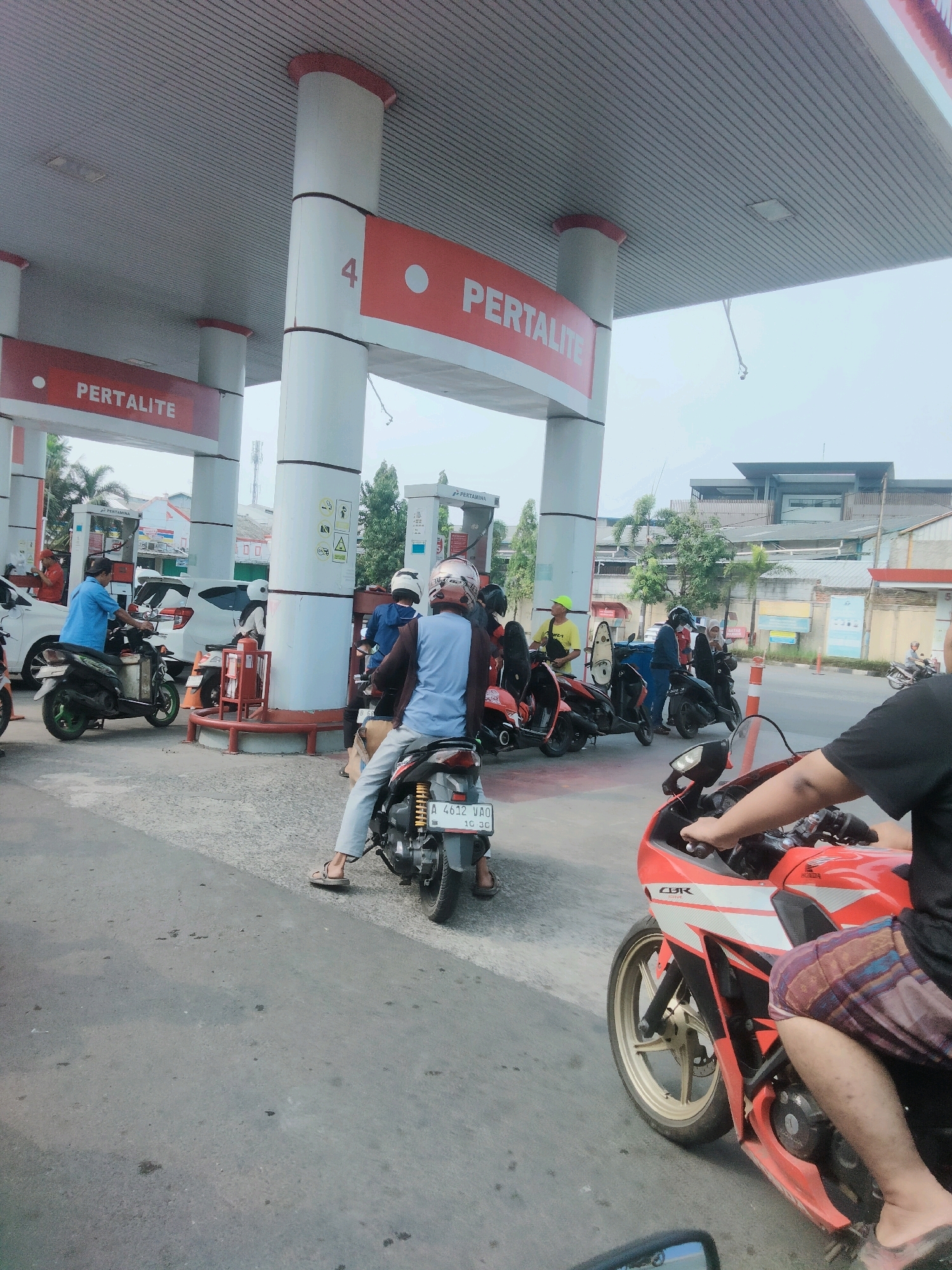 The image depicts a busy scene at a Pertamina gas station in Tangerang, Indonesia. Multiple people on motorcycles are waiting to refuel. In the foreground, a person in a black shirt and plaid shorts is seated on a red and white motorcycle, with their hands on the handlebars. To the left, another individual in a blue shirt is on a dark-colored motorcycle. Behind them, several other motorcycles and a white car are visible, suggesting a queue. The gas station features tall white pillars with red trim, displaying "PERTALITE" signage. The overall environment is an outdoor setting under a covered structure, typical of a gas station, with a bright, overcast sky.