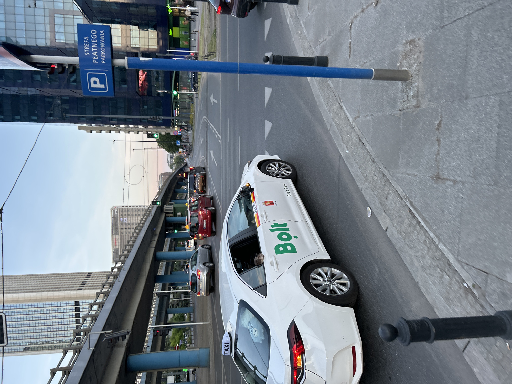 The image shows a street scene in Warsaw, Poland. A white Bolt taxi is positioned on the right side of the road, close to the curb. A blue parking sign, labeled "STREFA PLATNEGO PARKOWANIA", is visible, indicating paid parking. The background includes a bridge and several buildings. The sky is visible above the buildings. The street has traffic and lane markings. The photo is taken at a slightly tilted angle, and there is a black post in the bottom right corner.