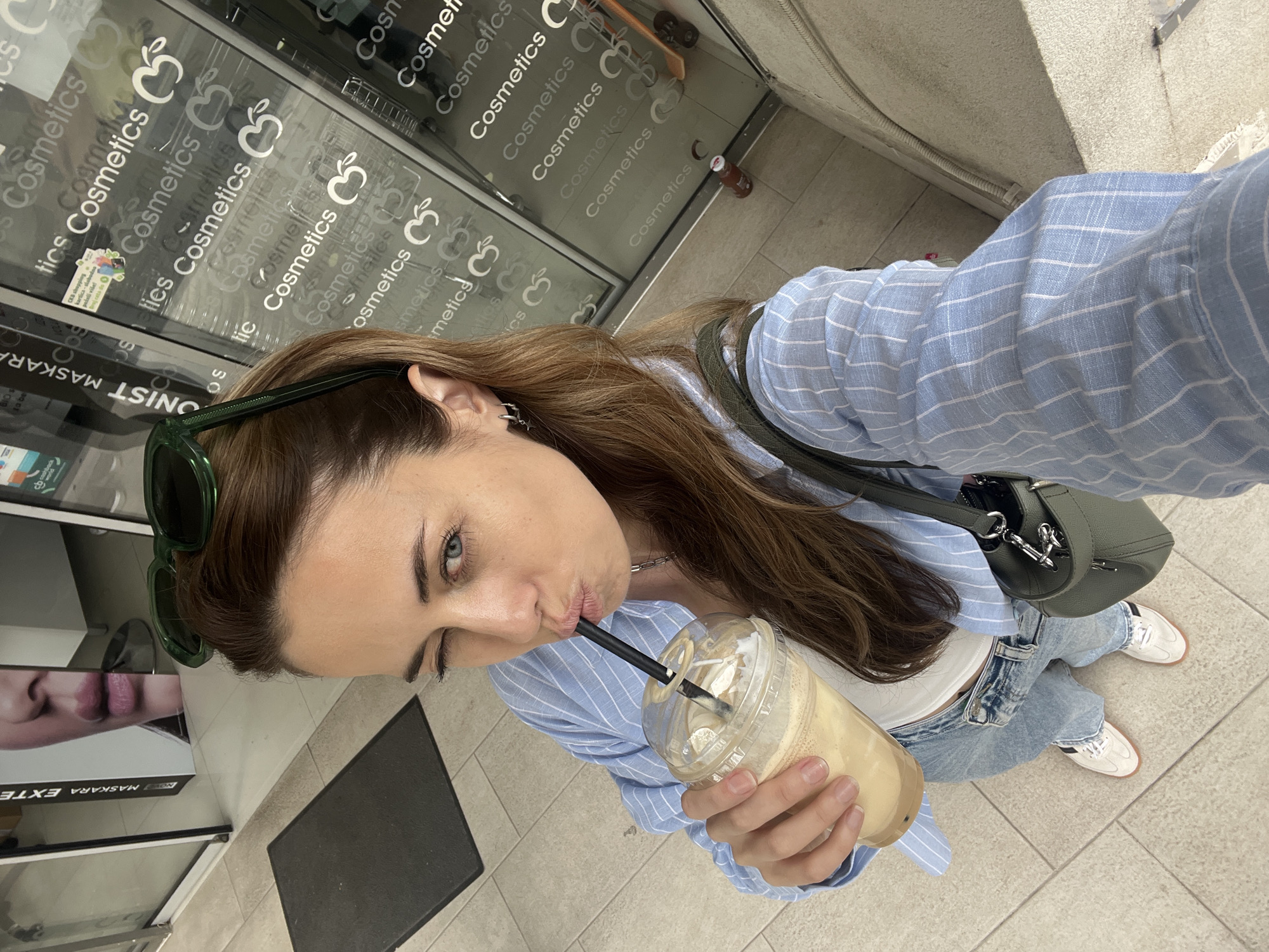 A woman is taking a selfie while winking and drinking from a plastic cup with a straw. She is wearing a light blue striped shirt, white top, light blue jeans, and white sneakers. She has sunglasses on her head and is wearing a green crossbody bag. She is standing in front of a store with the word "Cosmetics" written on the glass multiple times along with an apple logo. There is a black doormat to her left, and a poster with the word "MASKARA EXTE" is partially visible. Based on the text on the shop front window, it is located in the city of Bar, Montenegro.