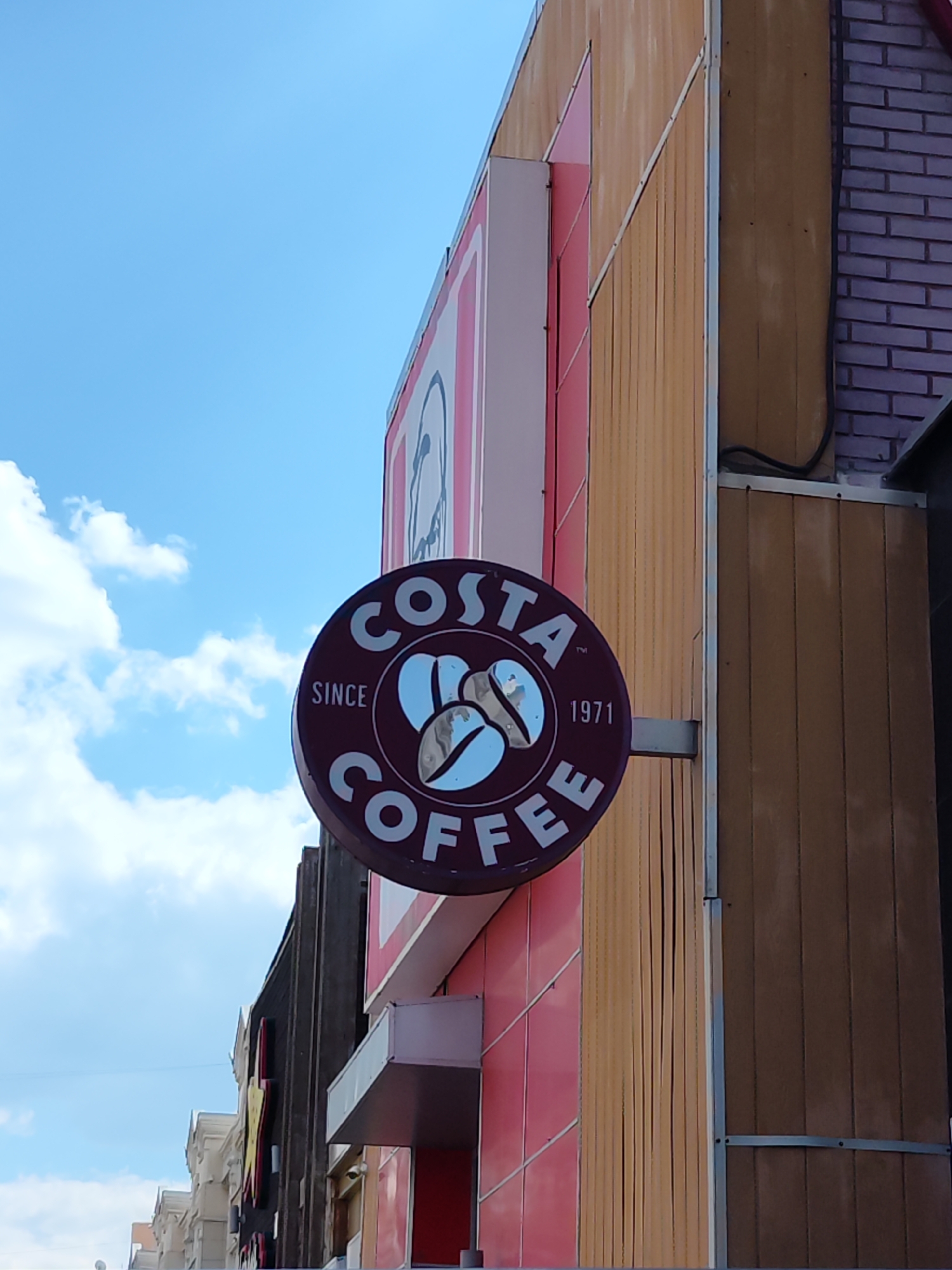 An exterior shot of a Costa Coffee shop with a circular sign. The sign is dark red with white lettering that reads "COSTA COFFEE" and "SINCE 1971". A stylized coffee bean logo is in the center of the sign. The shop's facade is a mix of vertical wood-like panels and pink tiled sections. The sky is blue with white clouds. The image appears to be taken from a low angle, looking up at the sign. Based on the architectural style and context, this Costa Coffee shop is located in Astana, Kazakhstan.