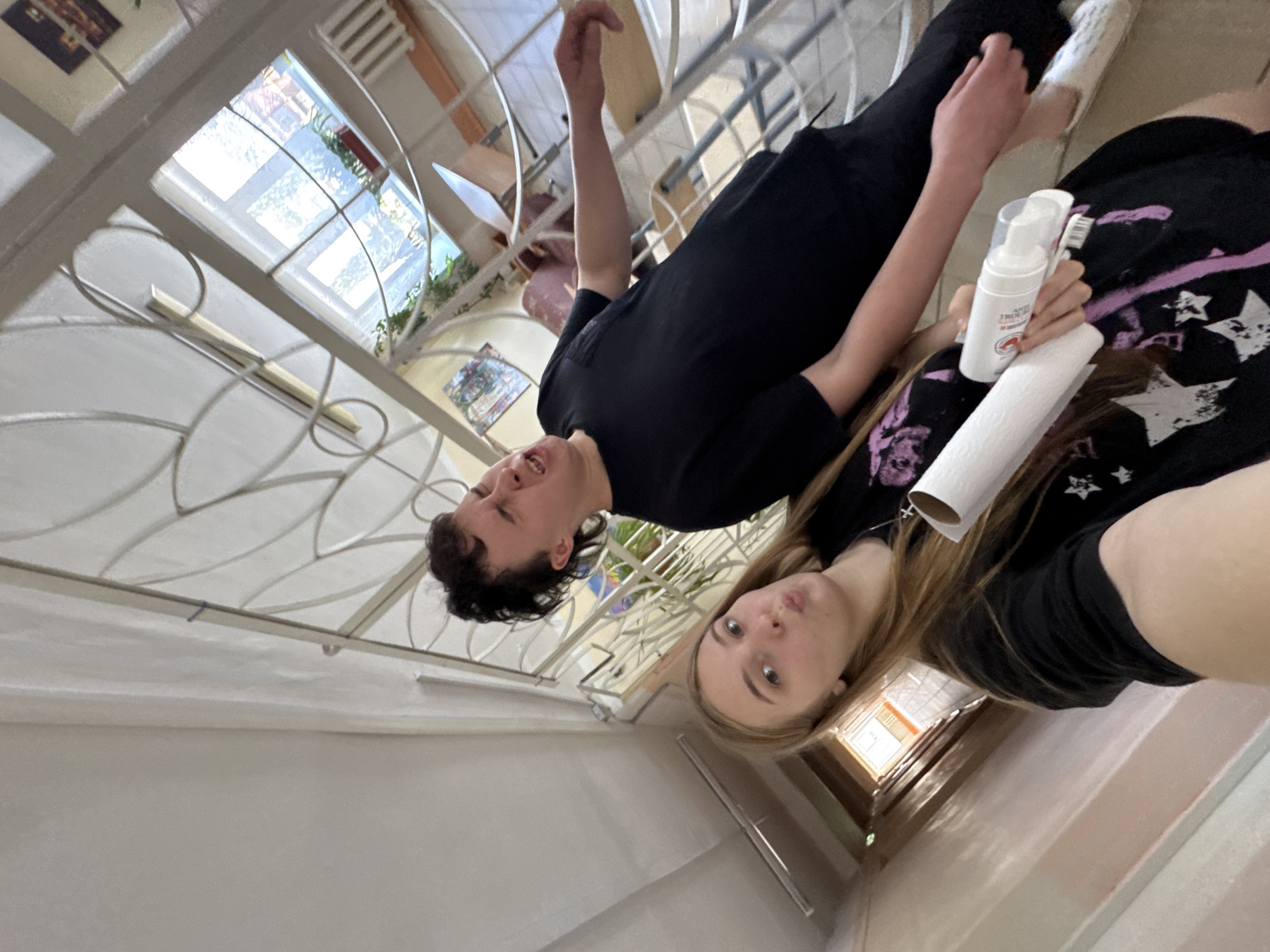 Two individuals, a young woman and a young man, pose for a selfie in what appears to be a school hallway. The young woman, in the foreground, has long blonde hair and is wearing a black t-shirt with a purple design. She is holding a roll of paper towels and a white bottle with a dispenser. The young man, standing behind and to the right of her, has dark, messy hair and is wearing a black t-shirt. He has his mouth open and appears to be making a funny face. The hallway is brightly lit, with a window visible in the background showing plants and sunlight. The walls are painted a light color, and there is a railing with decorative metalwork. The scene suggests a casual, informal moment, likely within an educational institution. This specific location cannot be identified within the city of Perm, Russia, based solely on the visual information provided.