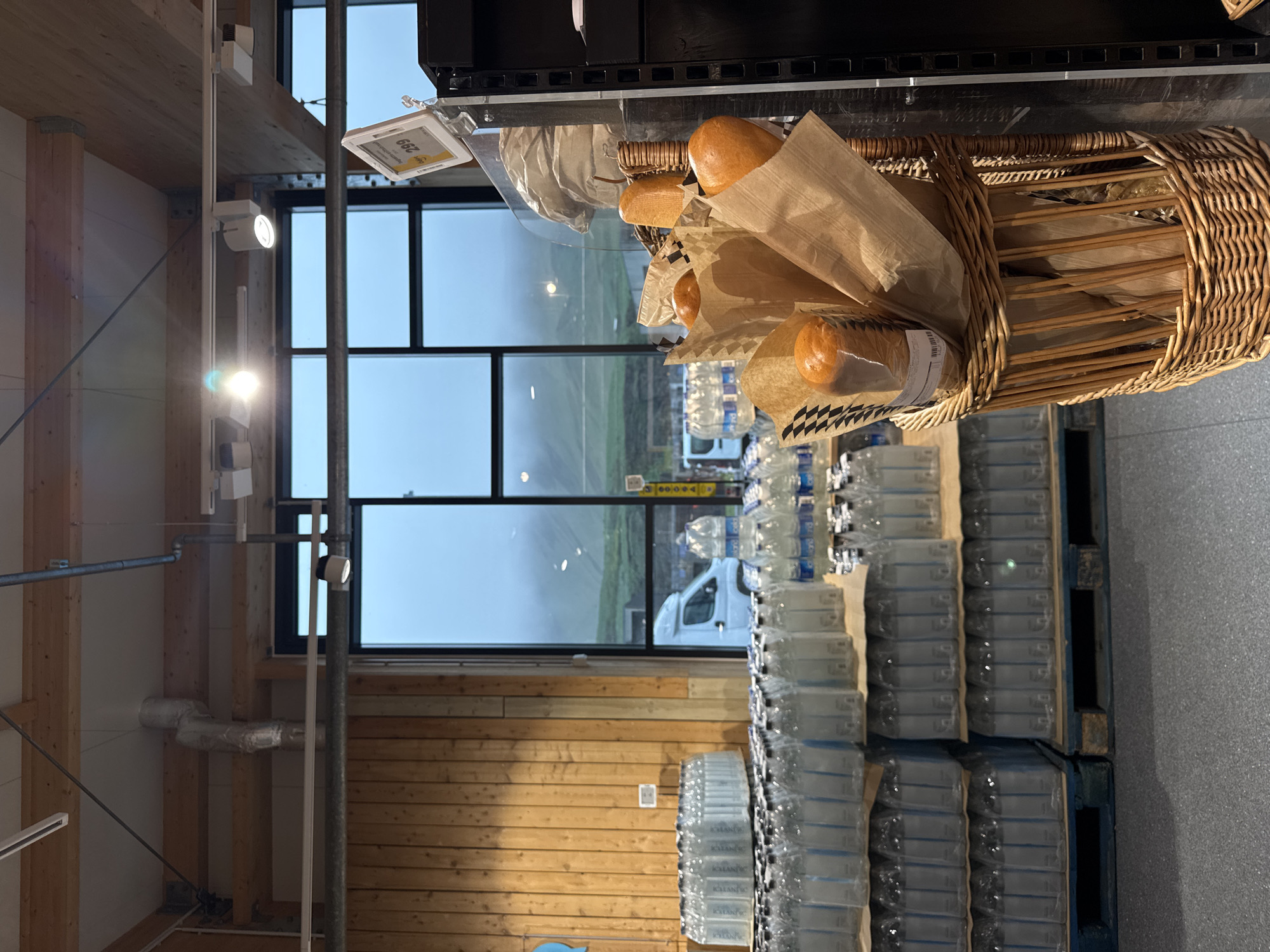 The image captures an interior scene at a shop in Vik, Iceland. It features a wicker basket filled with wrapped baguettes displayed above pallets stacked with bottled water. Through the window, the landscape outside is visible, revealing a foggy or misty environment. A white van is parked outside. The interior architectural elements include wooden beams and a series of rectangular windows. A price tag is visible near the baguettes.