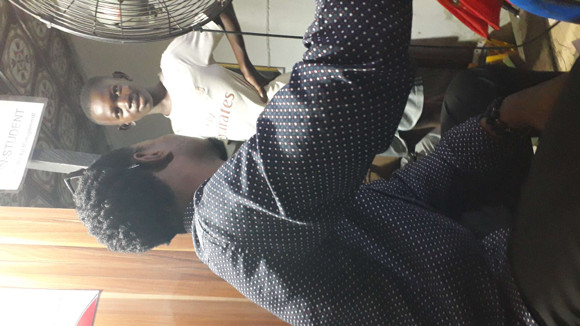 In Onitsha, Nigeria, two individuals are captured in a casual indoor setting, possibly a home or a small business. The primary subject, a young Black male with dark, curly hair and wearing a dark blue shirt with white polka dots, is shown from the back and side, looking towards the right of the frame. He has a faint beard and is wearing eyeglasses. To his right stands a younger Black male, facing the camera with a slight smile. He is wearing a white t-shirt with faded lettering. The background features a wall with a wooden texture on the left, a sign with "IN-STUDENT" visible at the top, and a large industrial fan on the right. The lighting appears to be dim, suggesting an evening or indoor environment with artificial light sources. The overall atmosphere is relaxed and informal, with the two individuals seemingly engaged in conversation or observing something together.