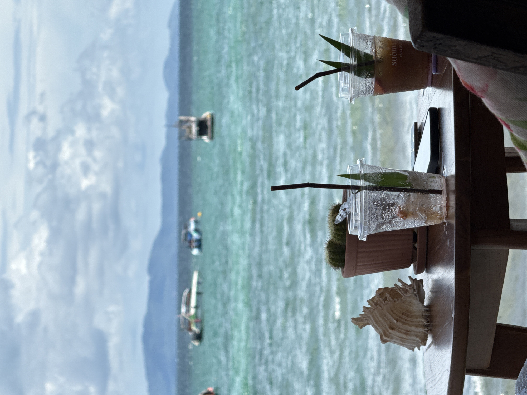 The image, taken in Pattaya City, Thailand, shows an outdoor table with two plastic cups containing drinks. One cup has a reddish-brown liquid and the word "subma" printed on its side; both have straws and green leaves as garnish. Next to the cups is a smartphone and a small potted cactus. A large seashell sits on the table's edge. In the background, the turquoise sea is visible with several small boats, and a cloudy sky above the distant coastline. The table appears to be part of an outdoor seating area, with a wooden chair partially visible in the foreground. The image is oriented vertically.
