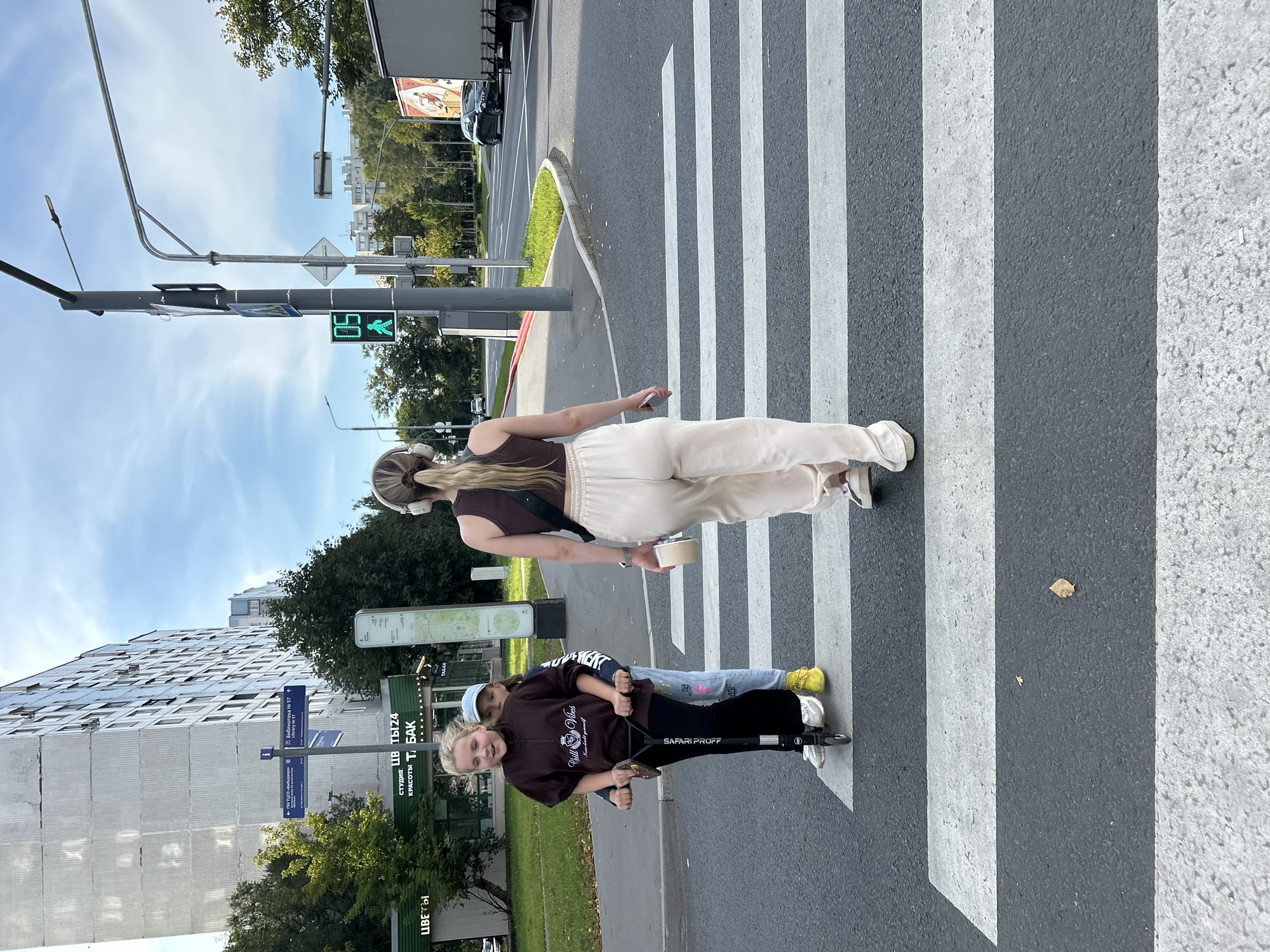 Two young girls are on a scooter crossing a pedestrian street in Moscow, Russia. The girl in front is wearing a brown sweatshirt and black pants, while the girl behind her is wearing a blue hat and a white t-shirt. A young woman with blonde hair and headphones is walking behind them. The street is a crosswalk with white stripes. A traffic light shows a green walking man symbol. In the background, there is a tall apartment building and trees. A truck is visible in the distance on the right side of the image.