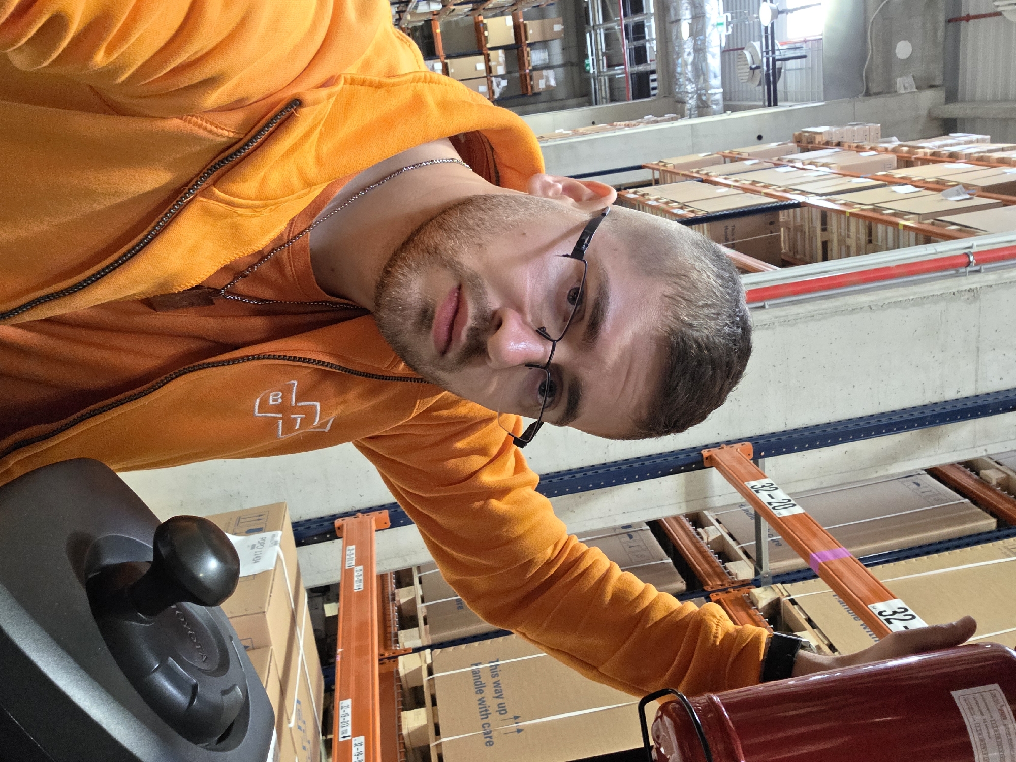 A man with a beard and glasses, wearing an orange hooded sweatshirt, is in a warehouse in Plovdiv, Bulgaria. He appears to be operating a forklift, based on the presence of a joystick control and the warehouse shelving with boxes. He has one hand on a red fire extinguisher. The warehouse has concrete walls and ceilings with metal shelving. The shelves are stocked with cardboard boxes.