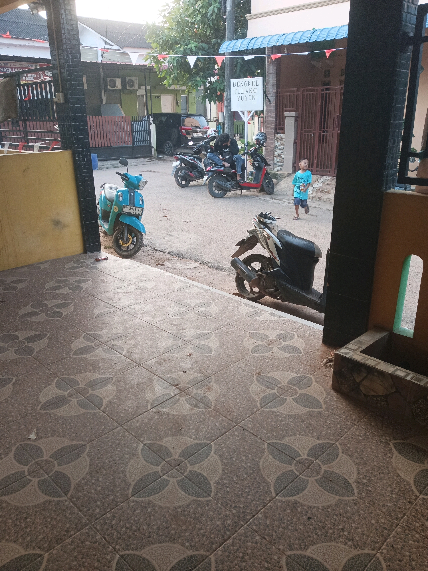 In Batam, Indonesia, a street scene unfolds with multiple motorcycles parked and in use. In the foreground, a light blue scooter with license plate BP 5978 EJ is parked. Further down the street, a black and white scooter is also parked. On the right side of the image, a young boy in a blue t-shirt and shorts walks away from the viewer. Behind him, two individuals are seated on a red motorcycle, and another red motorcycle is parked nearby. A white sign with black text that reads "BENGKEL TULANG YUYUN" is visible in the background. The environment suggests a residential or commercial area with buildings and fences.