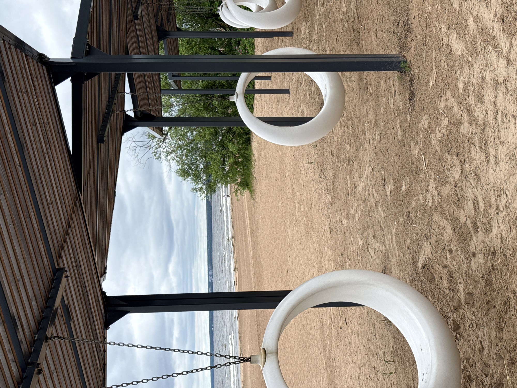 The image depicts a beach scene in Sestroretsk, Russia, featuring an outdoor seating area with a slatted wooden roof supported by black metal pillars. Several white, circular swings, resembling large donuts, hang from chains suspended from the roof structure. The swings are positioned over a sandy beach, with the Gulf of Finland visible in the background. The water is choppy, and the sky is overcast. There are a few distant figures walking on the beach, and some greenery, likely trees and bushes, are present behind the beach. The sand shows some footprints and traces of tire tracks. The overall atmosphere is serene and suggests a public recreational space.