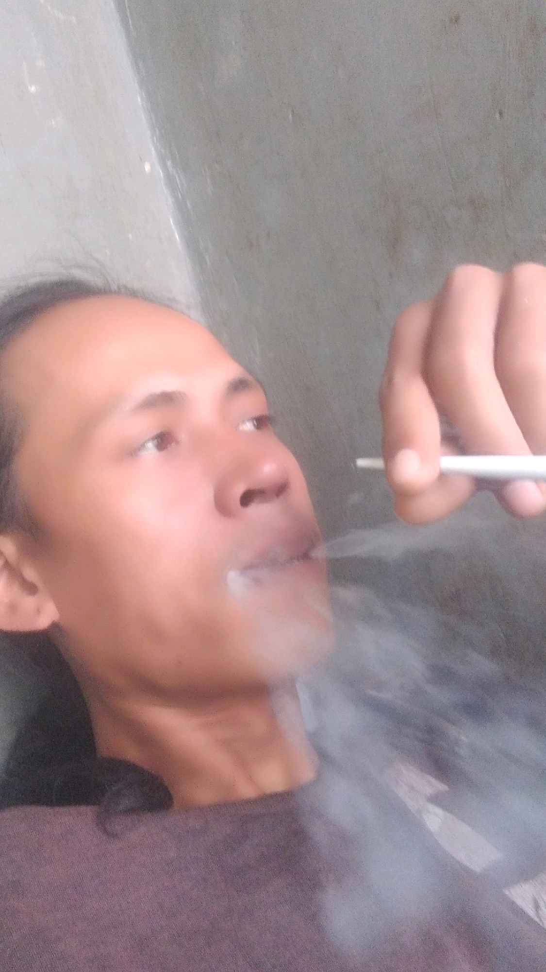 A close-up, high-angle, medium shot shows a person exhaling smoke from a cigarette, held to their mouth by their right hand. The person's face is turned to the right and slightly upward, with their eyes looking away. They are wearing a dark purple t-shirt. The background consists of a rough, grey wall. The scene appears to be outdoors in Rahong, Indonesia.