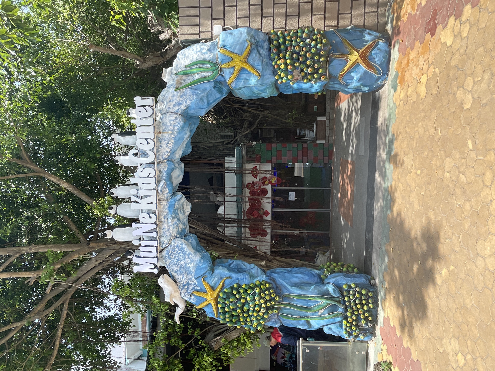 The image shows the entrance to "Mui Ne Kids Center" in Mũi Né, Vietnam. The entrance is an archway decorated with a blue, wave-like design, seaweed, starfish, and clusters of green and yellow spheres resembling fruit or coral. Five white penguin figurines are perched on top of the arch. A white dolphin sculpture is positioned on the left side of the arch. The ground in front of the entrance is paved with interlocking hexagonal tiles in shades of red, yellow, and green. Behind the archway, a building with glass doors is visible, with hanging red decorations inside. To the left of the entrance, a person wearing a red jacket and a green cap is partially visible. The scene is set outdoors, with large trees providing shade overhead. The overall impression is a playful and inviting entrance to a children's center, likely located in a tropical or coastal area given the marine-themed decorations and lush greenery.