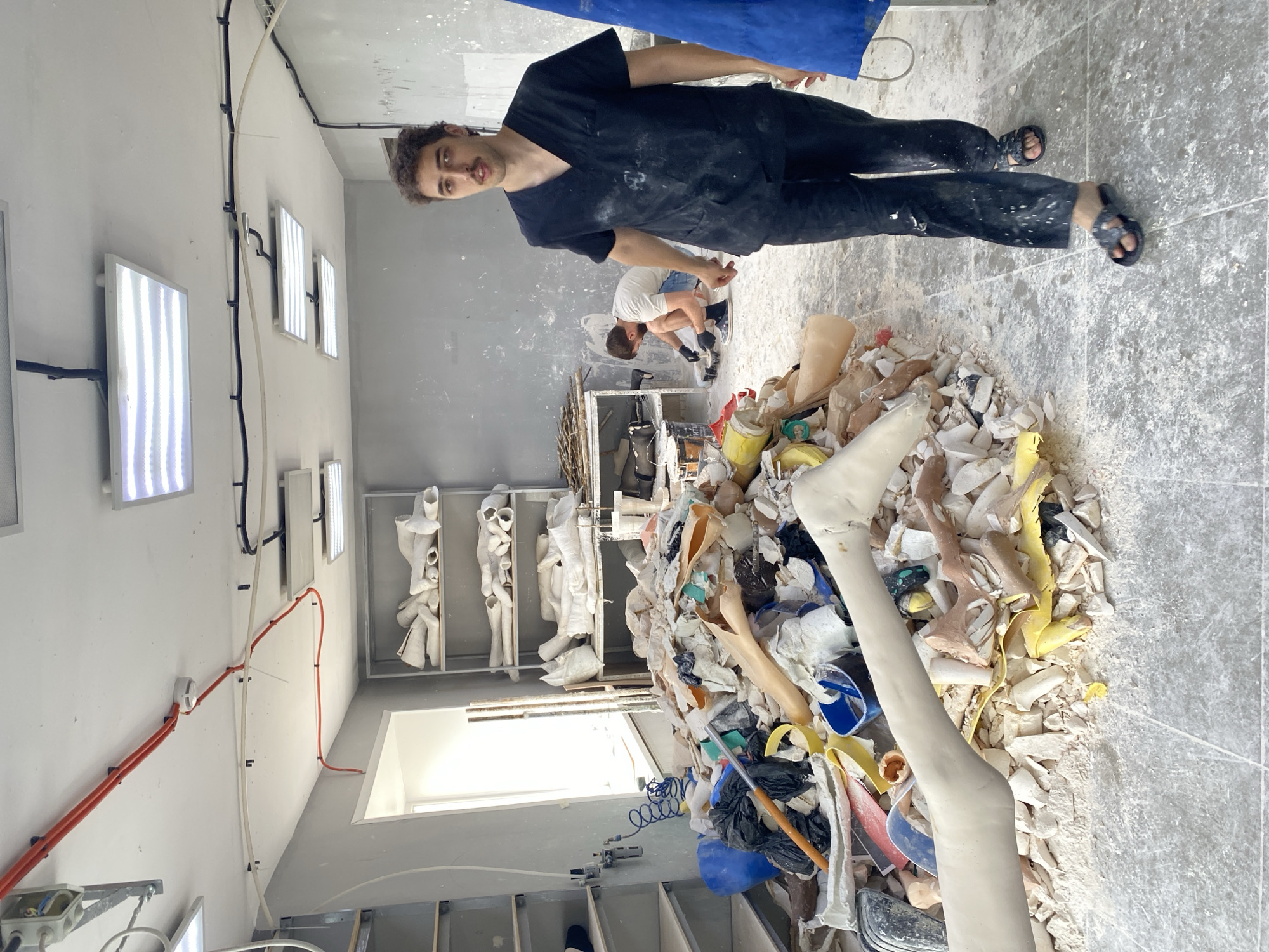 A man in dark scrubs walks past a large pile of discarded prosthetic limbs and workshop materials. Behind him, another man kneels and works on a small item. Shelves filled with white prosthetic parts line the back wall of a cluttered, brightly lit workshop. The floor and walls are dusty and stained, suggesting an active and messy workspace. Electrical conduits and light fixtures are visible on the ceiling. The scene appears to be an indoor workshop or laboratory.