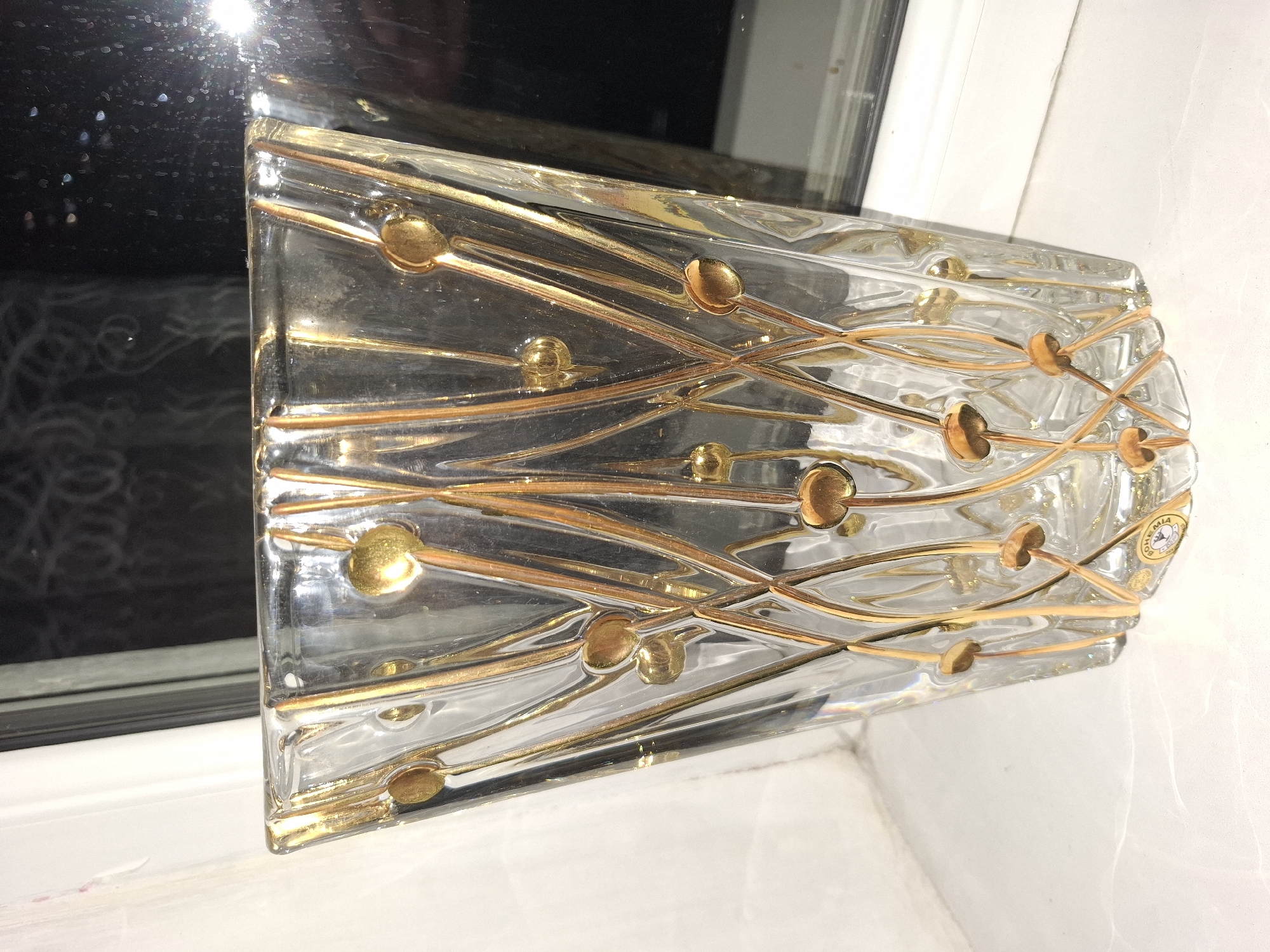 The image shows a clear glass vase with a decorative gold pattern. The vase is trapezoidal in shape, wider at the top than the bottom, and features thin gold lines that form an abstract design with small gold circles at intervals along the lines. The vase appears to be sitting on a white surface, possibly a windowsill, with a dark background visible behind it, suggesting it is near a window or reflective surface. A portion of a sticker with "BOHEMIA" is visible on the lower part of the vase. The image does not provide any information about the city of Almaty, Kazakhstan, or its location within the city.