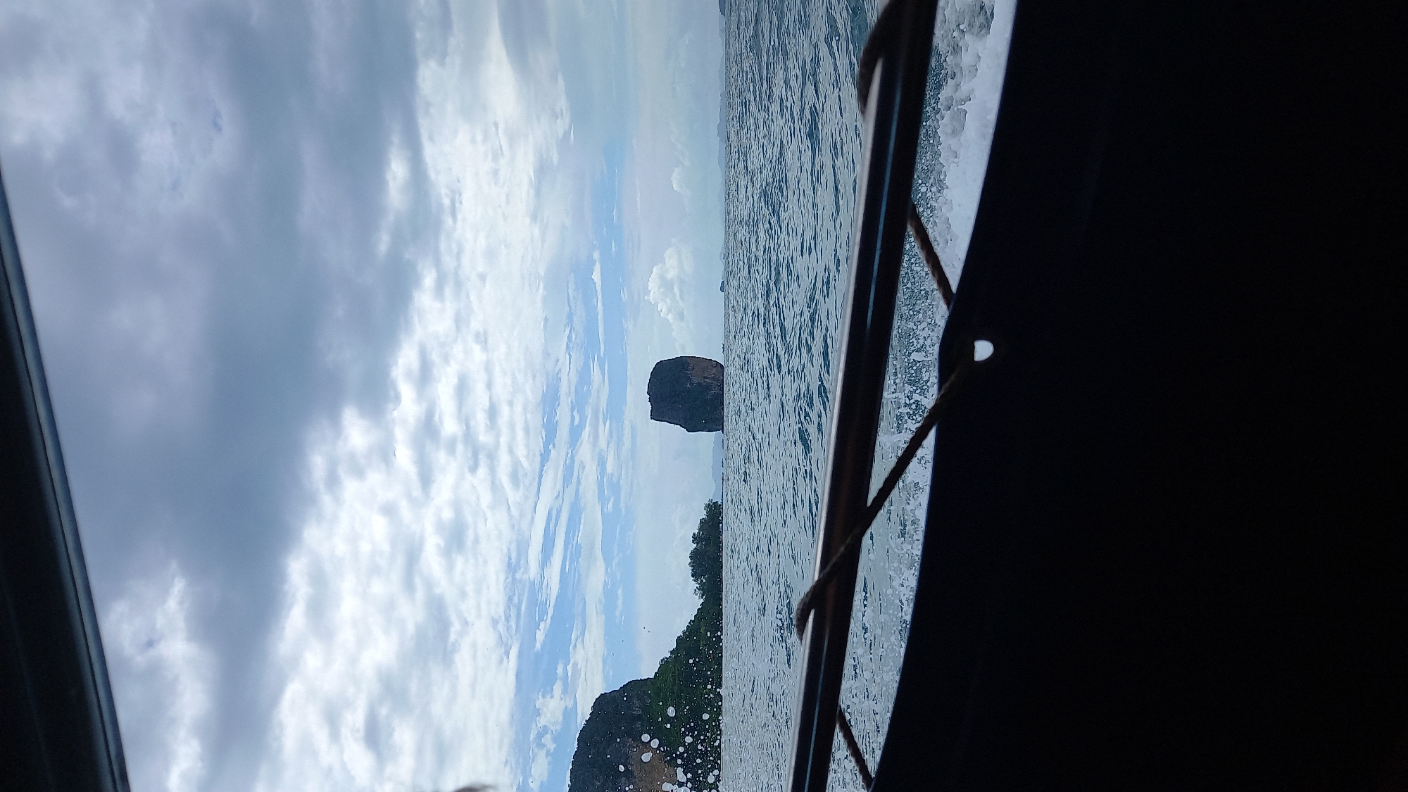 From the vantage point inside a moving boat, the frame captures a seascape off the coast of Ao Nang, Thailand. The ocean stretches out, with visible wave action caused by the boat's movement. A distinctive limestone karst formation rises from the water, a characteristic feature of the Ao Nang landscape. Landmasses covered with dense tropical vegetation are also visible along the horizon. The sky is overcast with varied cloud formations, creating a diffused light. The frame's foreground is dominated by elements of the boat's structure, including metal railings and shadowed interior sections.