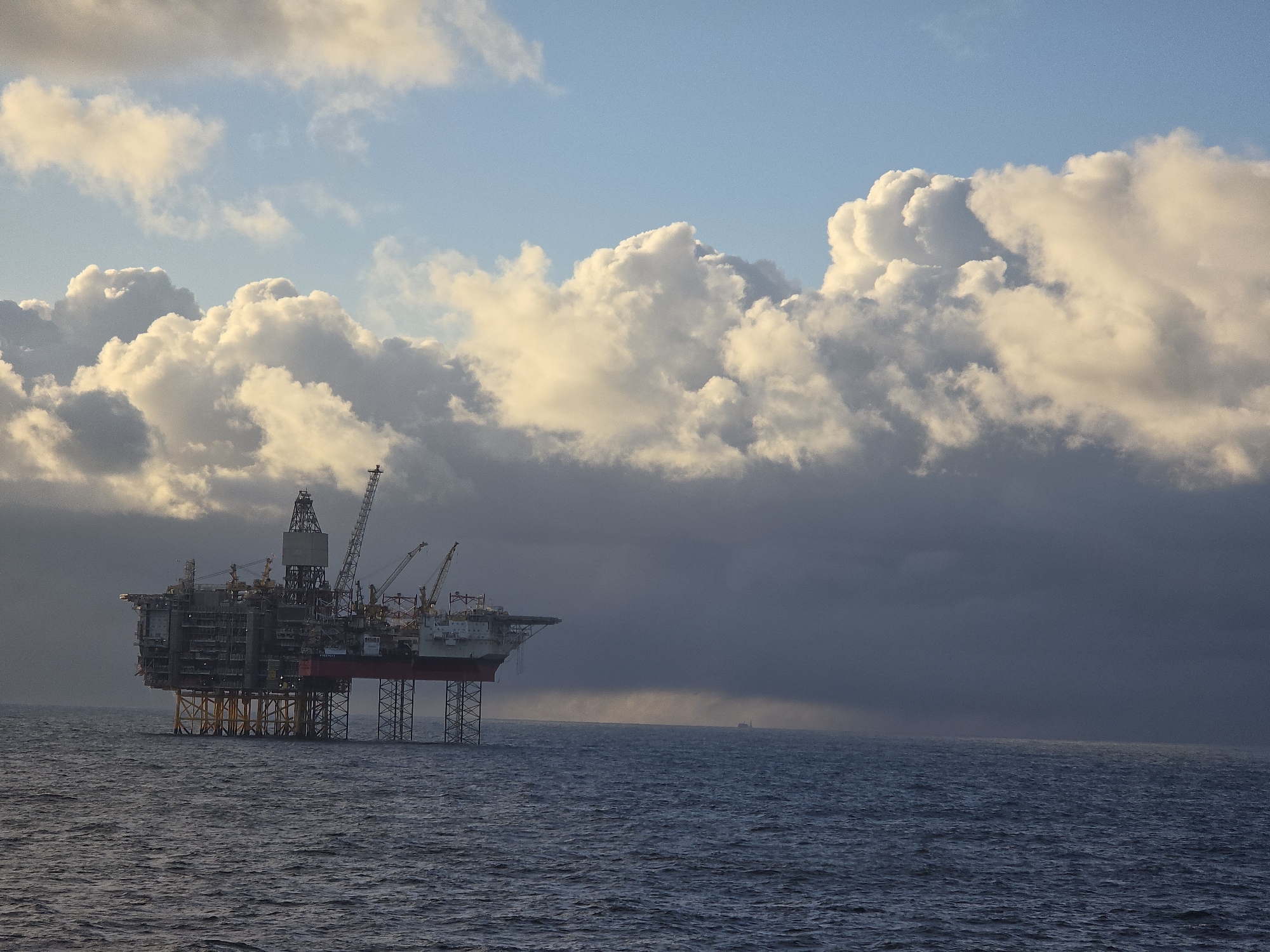 The image depicts an offshore oil platform standing in the ocean under a partly cloudy sky. The platform is a complex structure with multiple levels, cranes, and drilling equipment. The lower section of the platform is supported by a lattice framework extending down into the water. The sea is dark and choppy, with a distant outline of a small boat visible on the horizon. Rain is seen in the distance. The sky is a mixture of clear blue and cumulus clouds. The location is Unknown, Unknown.